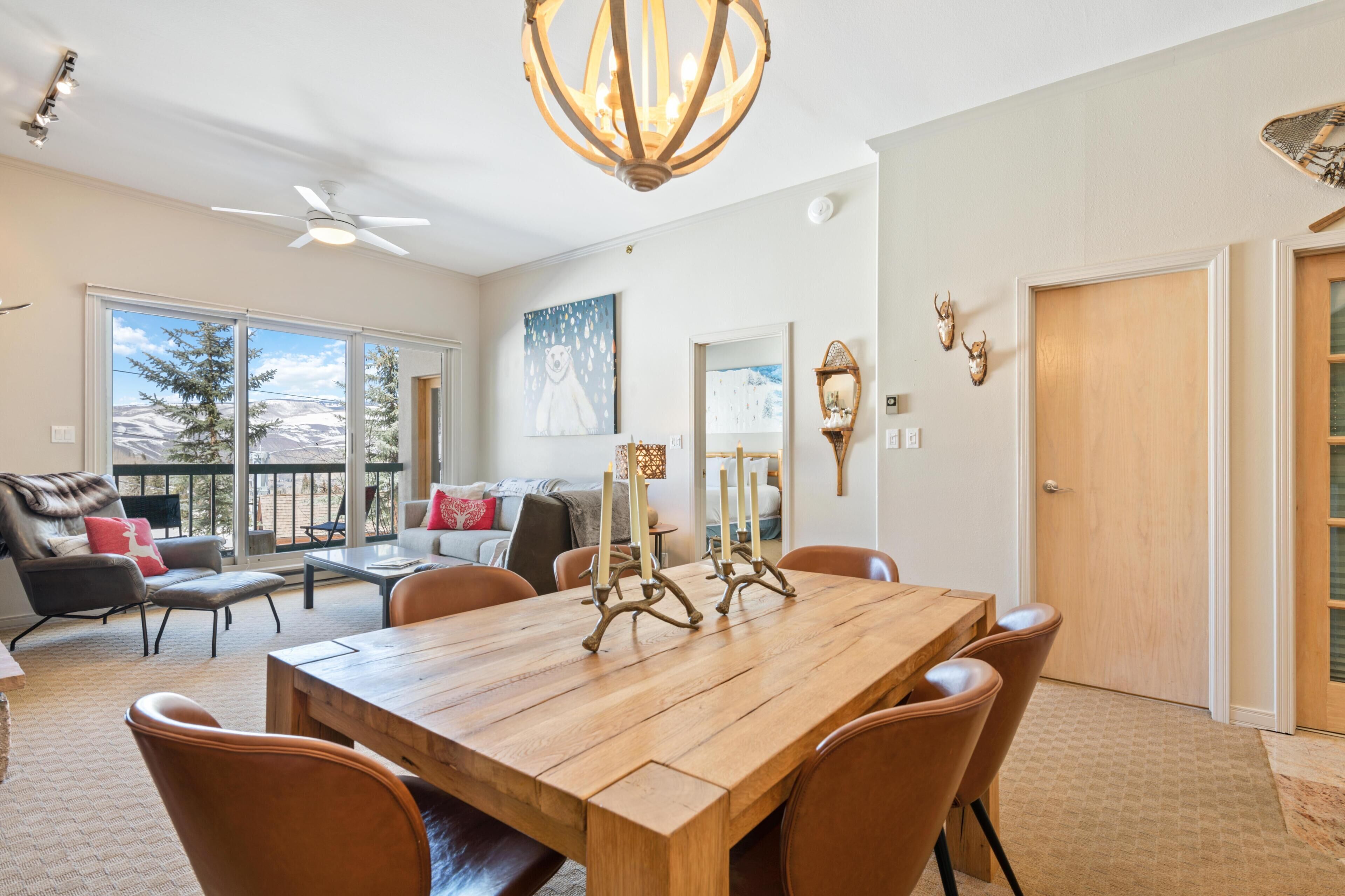 A spacious living room with a large wooden dining table, comfortable seating, a ceiling fan, and a view through large sliding glass doors.