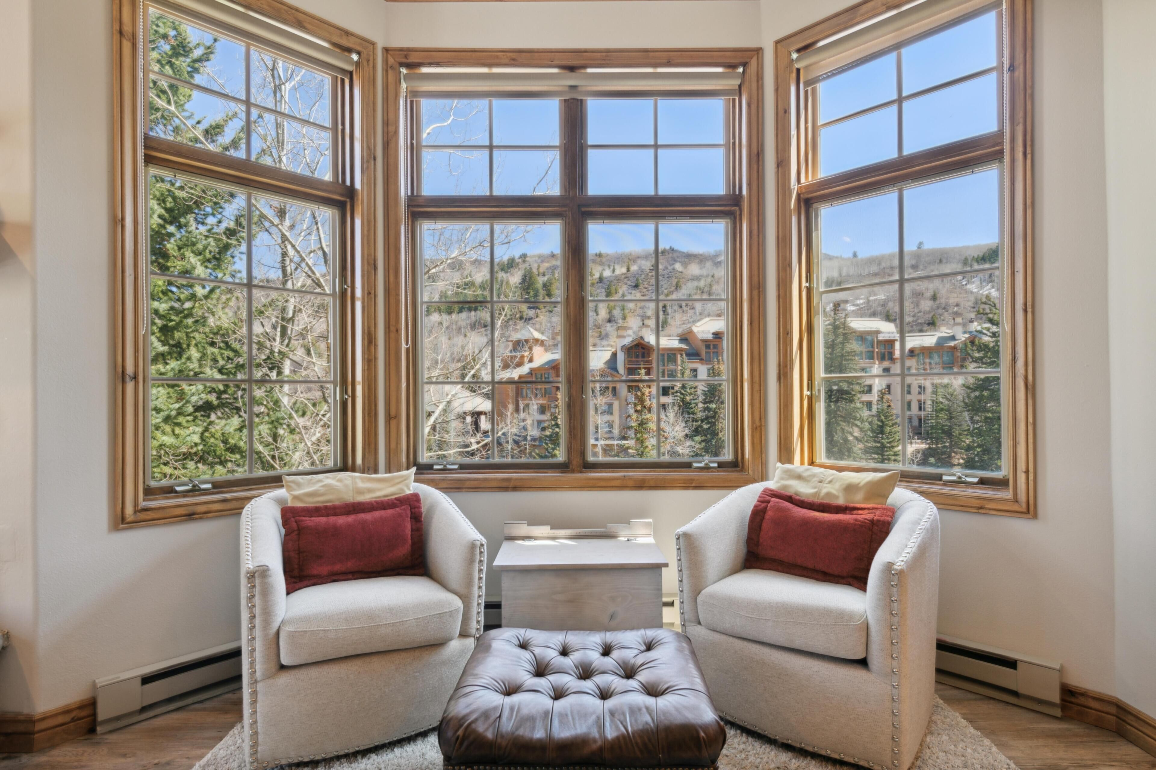 A room with a large bay window, two armchairs, a small table, and a leather ottoman.