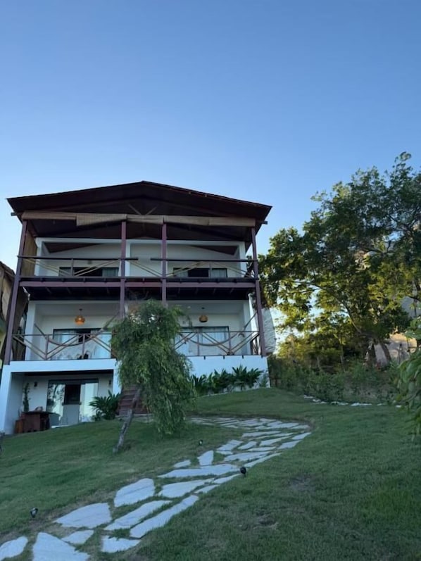 Un edificio de dos pisos con techo plano, un balcón en el nivel superior y un camino de piedra que conduce a la entrada.