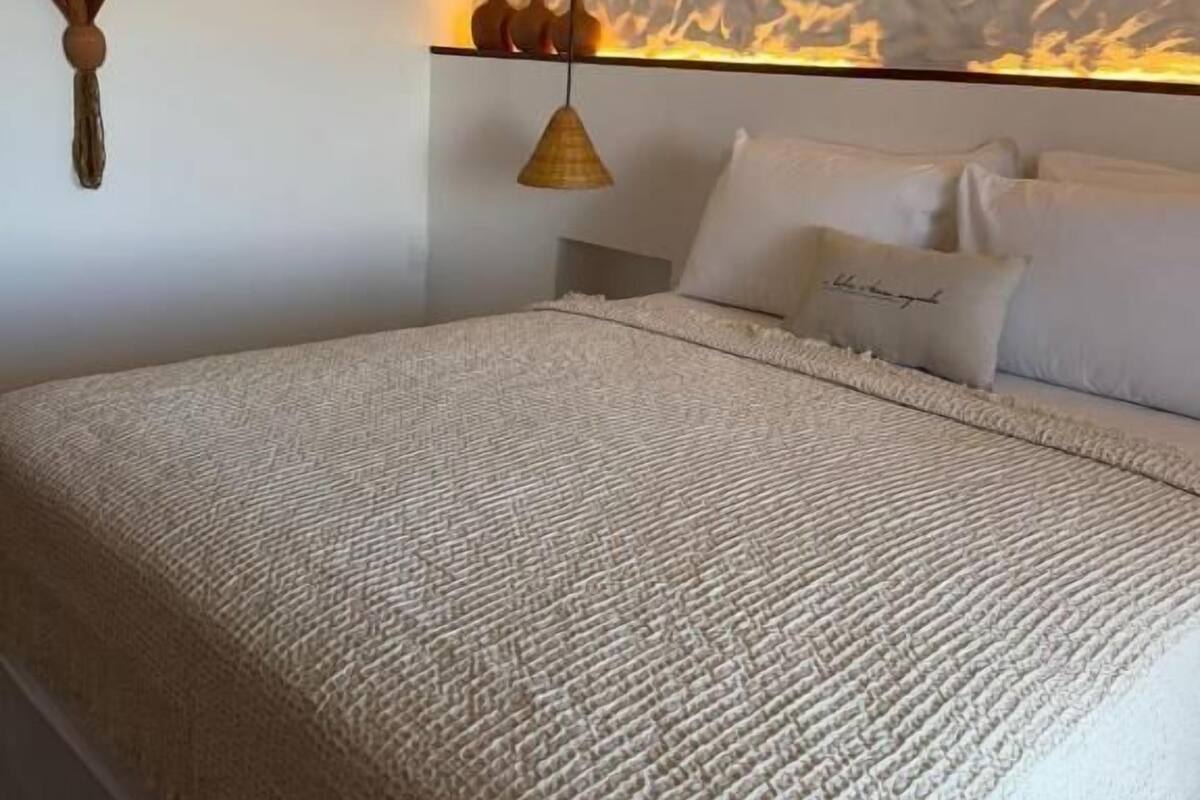 Un dormitorio con cama, ropa de cama blanca, una lámpara colgante y un equipo de aire acondicionado en la pared.