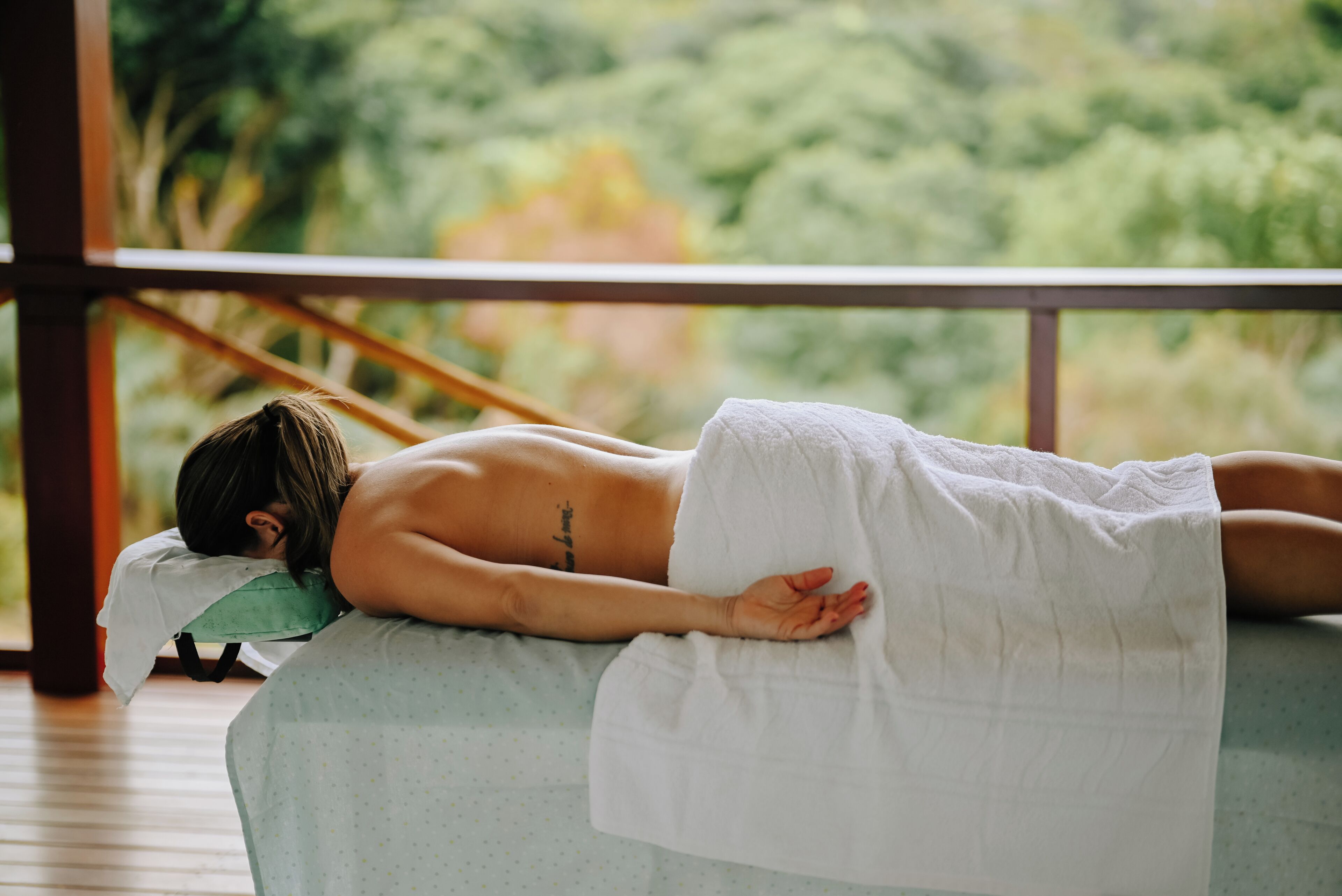 Une personne allongée sur une table de massage, recouverte d’une serviette sur la partie inférieure du corps, dans une pièce d’où l’on peut admirer la verdure à l’extérieur.