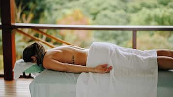 Une personne allongée sur une table de massage, recouverte d’une serviette sur la partie inférieure du corps, dans une pièce d’où l’on peut admirer la verdure à l’extérieur.