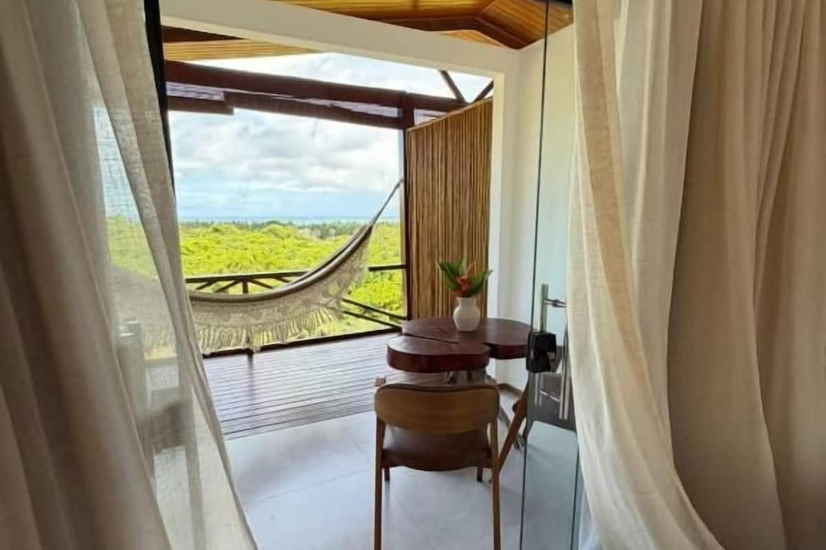 Una habitación con un ventanal grande, una hamaca, una silla de madera y una mesita con un florero.