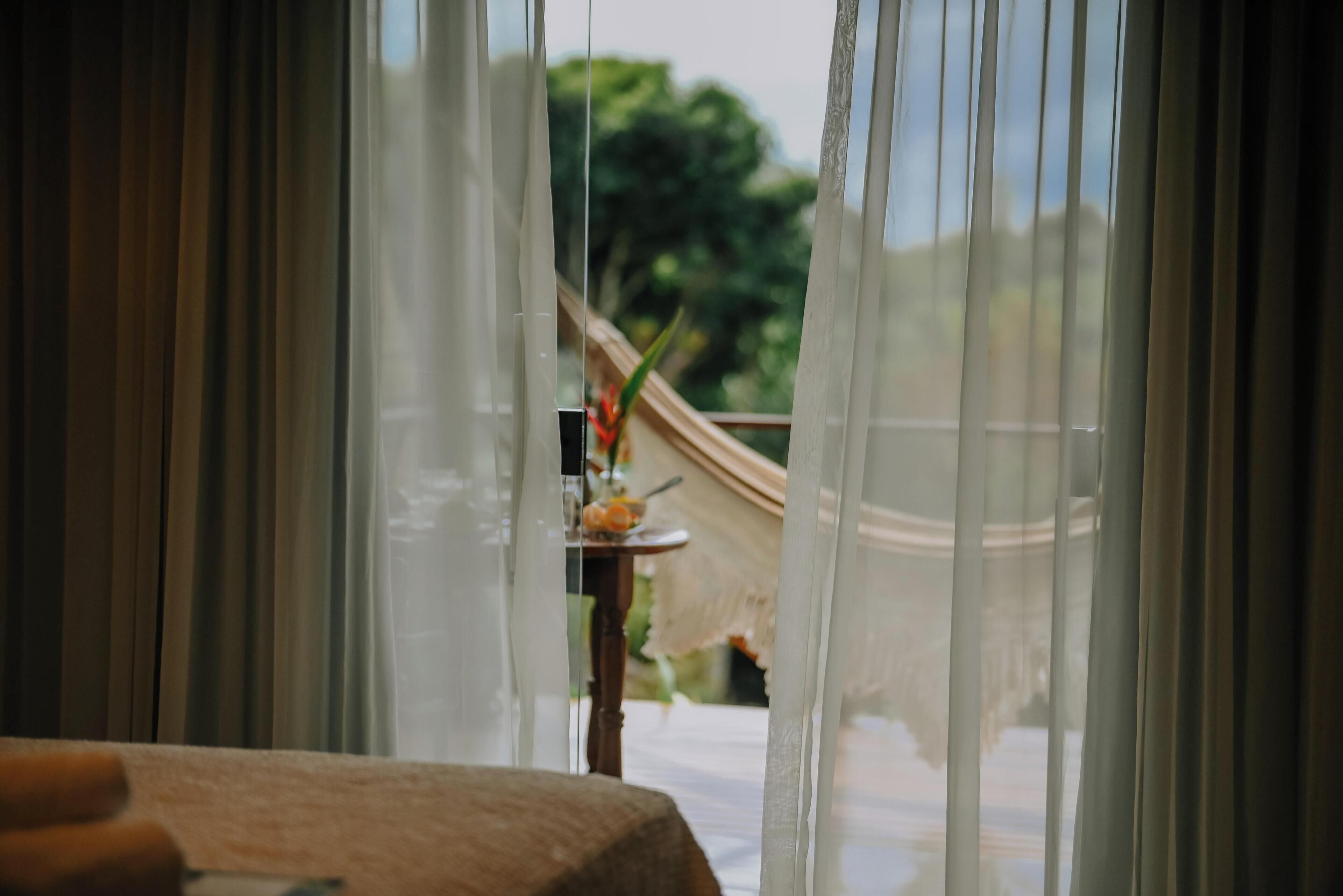 A room with a table and a view through sheer curtains to an outdoor area with greenery.