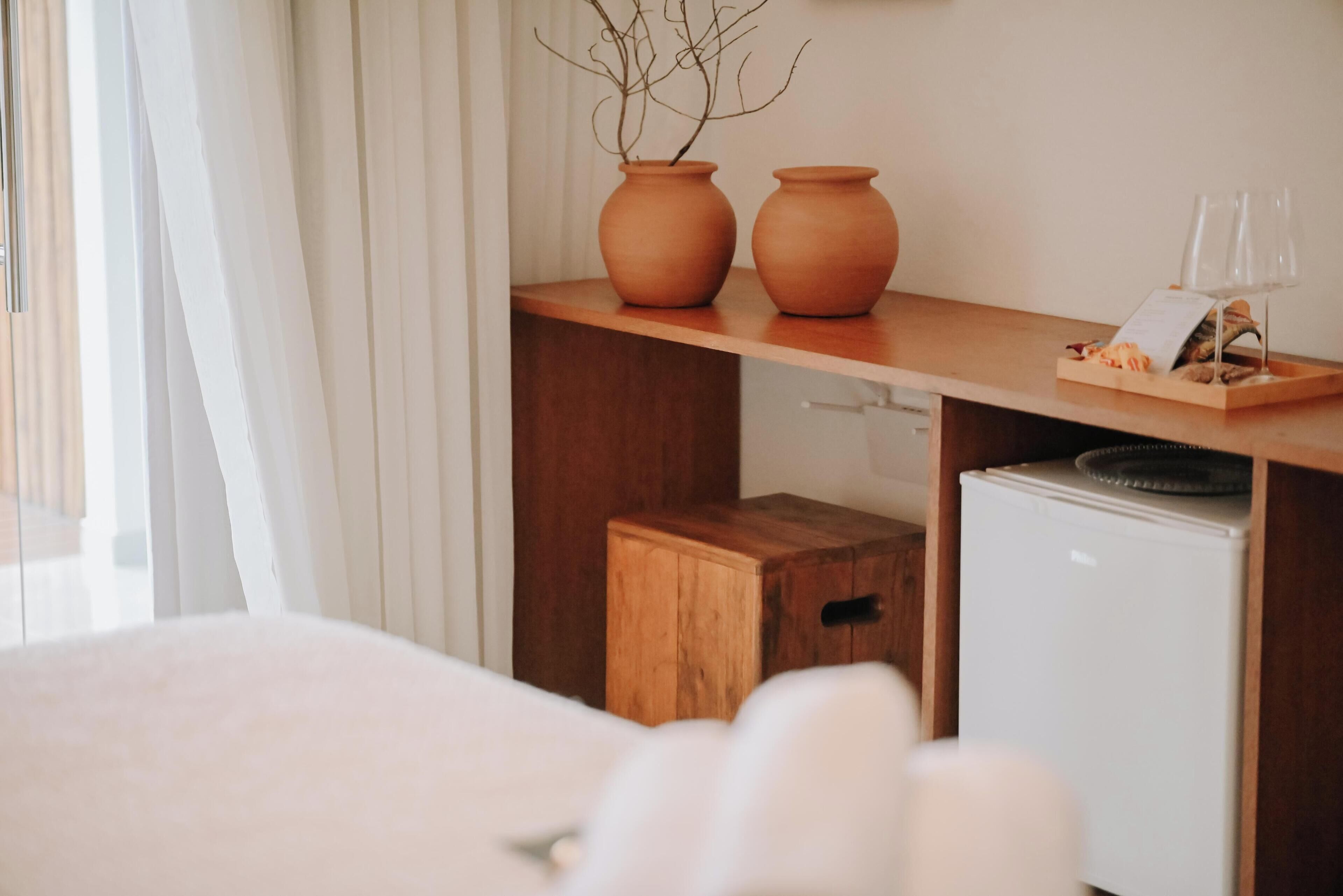 Una habitación de hotel con una cama, una mesita de noche de madera con un florero y vasos, y un refrigerador pequeño.
