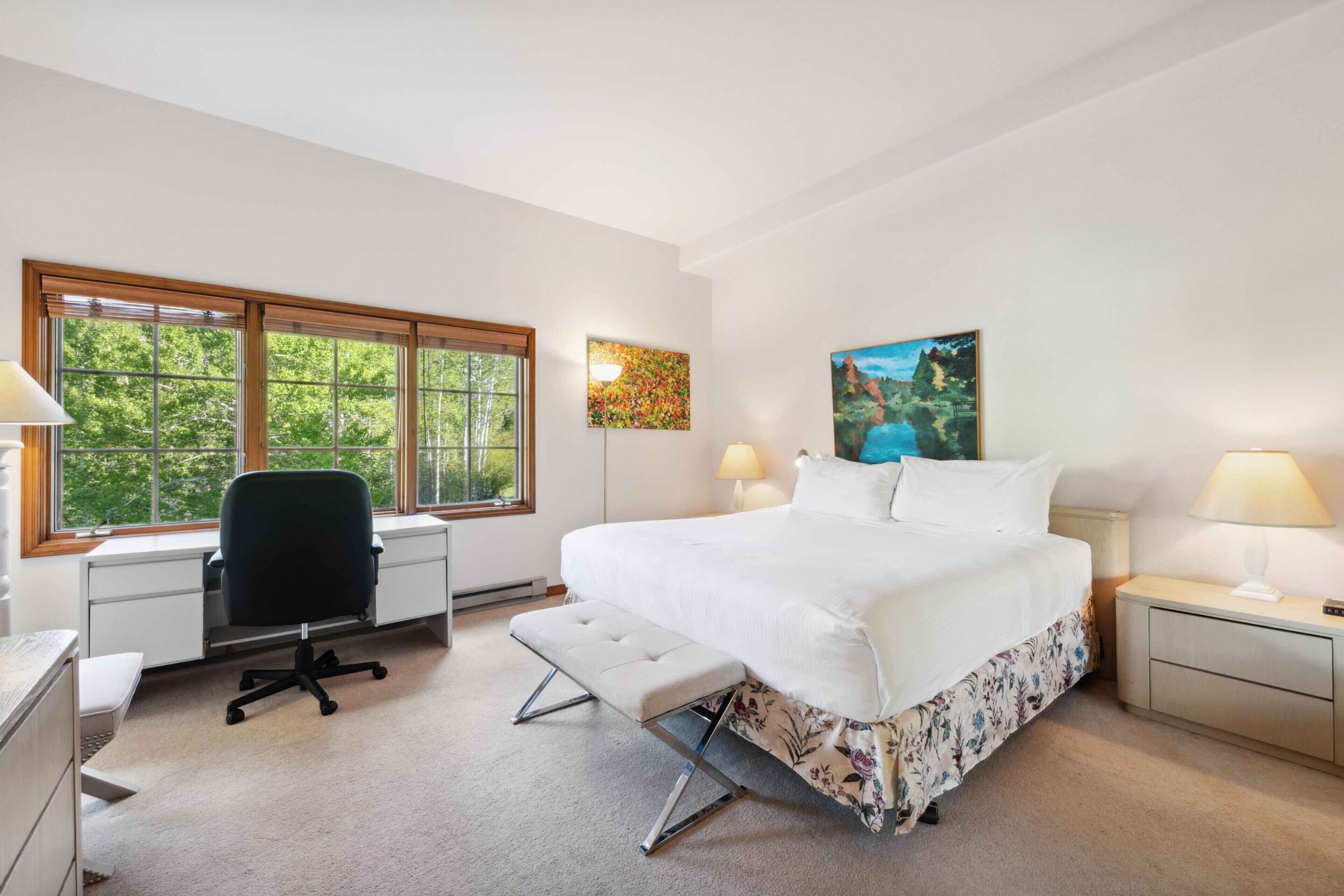 Quarto com cama, escrivaninha, cadeira, banco, mesas de cabeceira e obras de arte.
