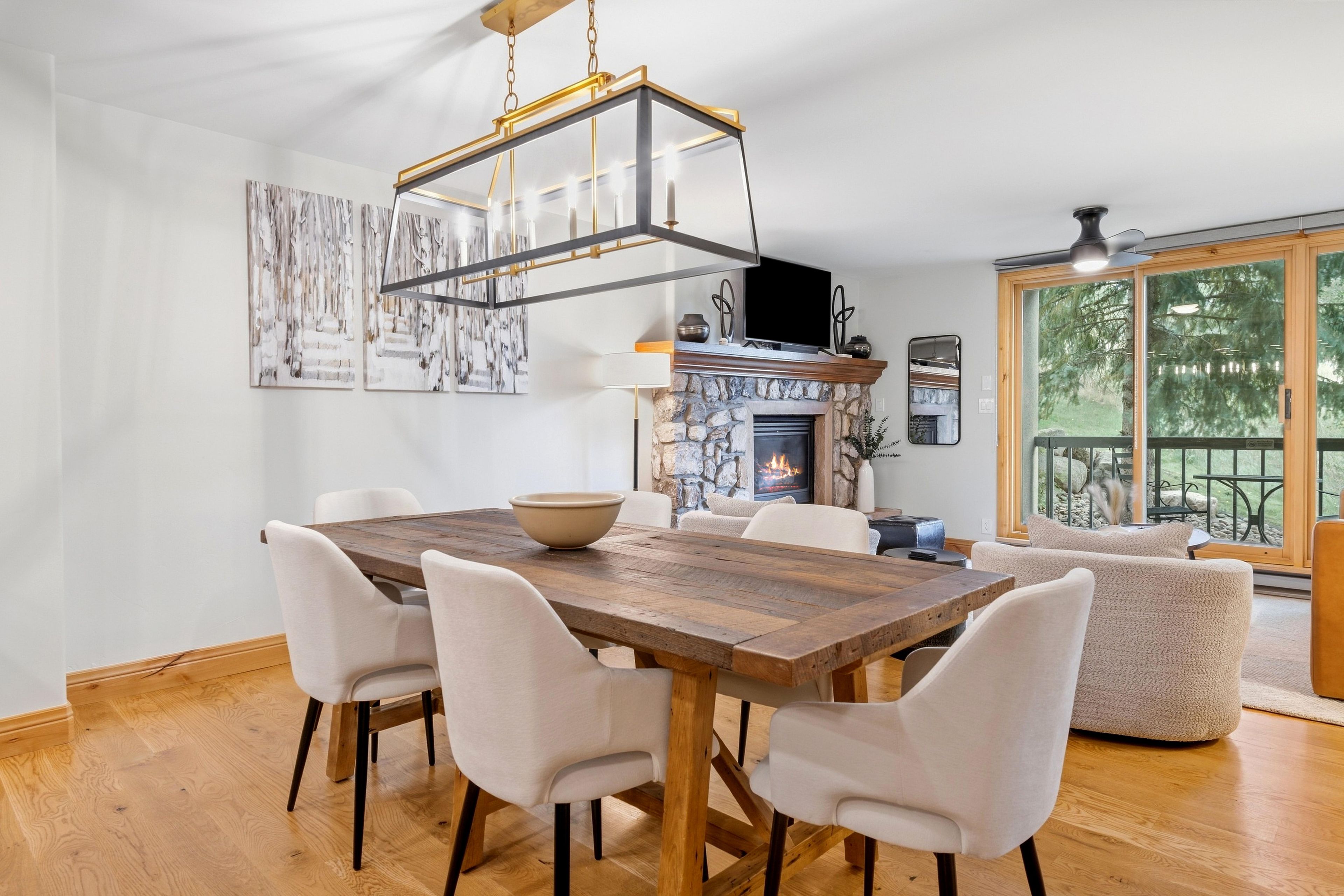 Un comedor con mesa de madera y sillas blancas, chimenea de piedra y un ventanal con vistas a árboles.