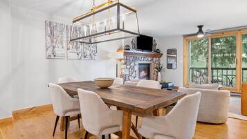 Un comedor con mesa de madera y sillas blancas, chimenea de piedra y un ventanal con vistas a árboles.