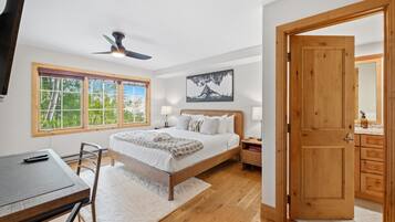 Un dormitorio con cama, mesitas de noche, escritorio, silla, ventilador de techo, ventana con vista a árboles y puerta que da a un baño.