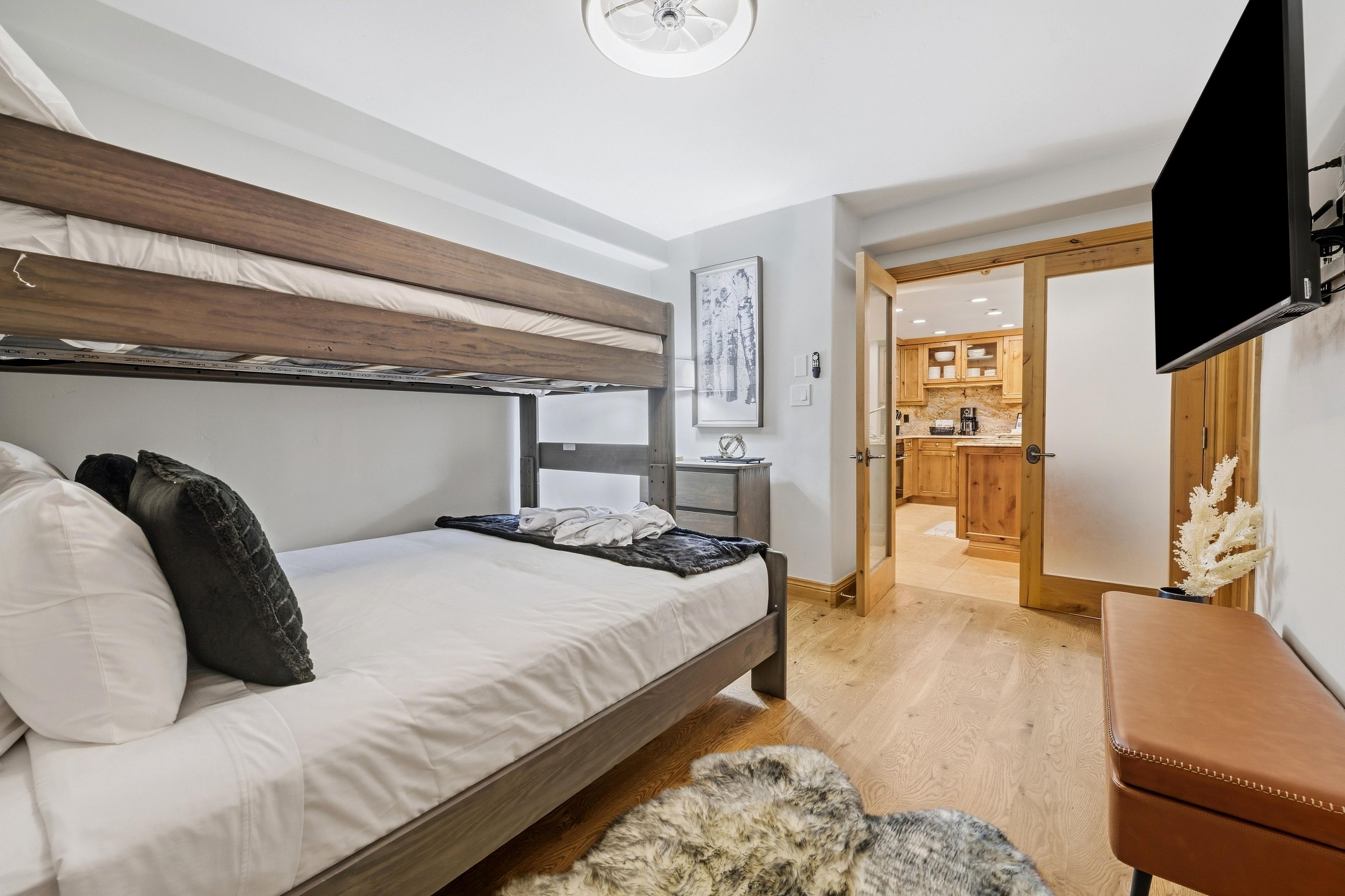A bedroom with a bunk bed, a desk, a chair, a television, and a view into the kitchen.