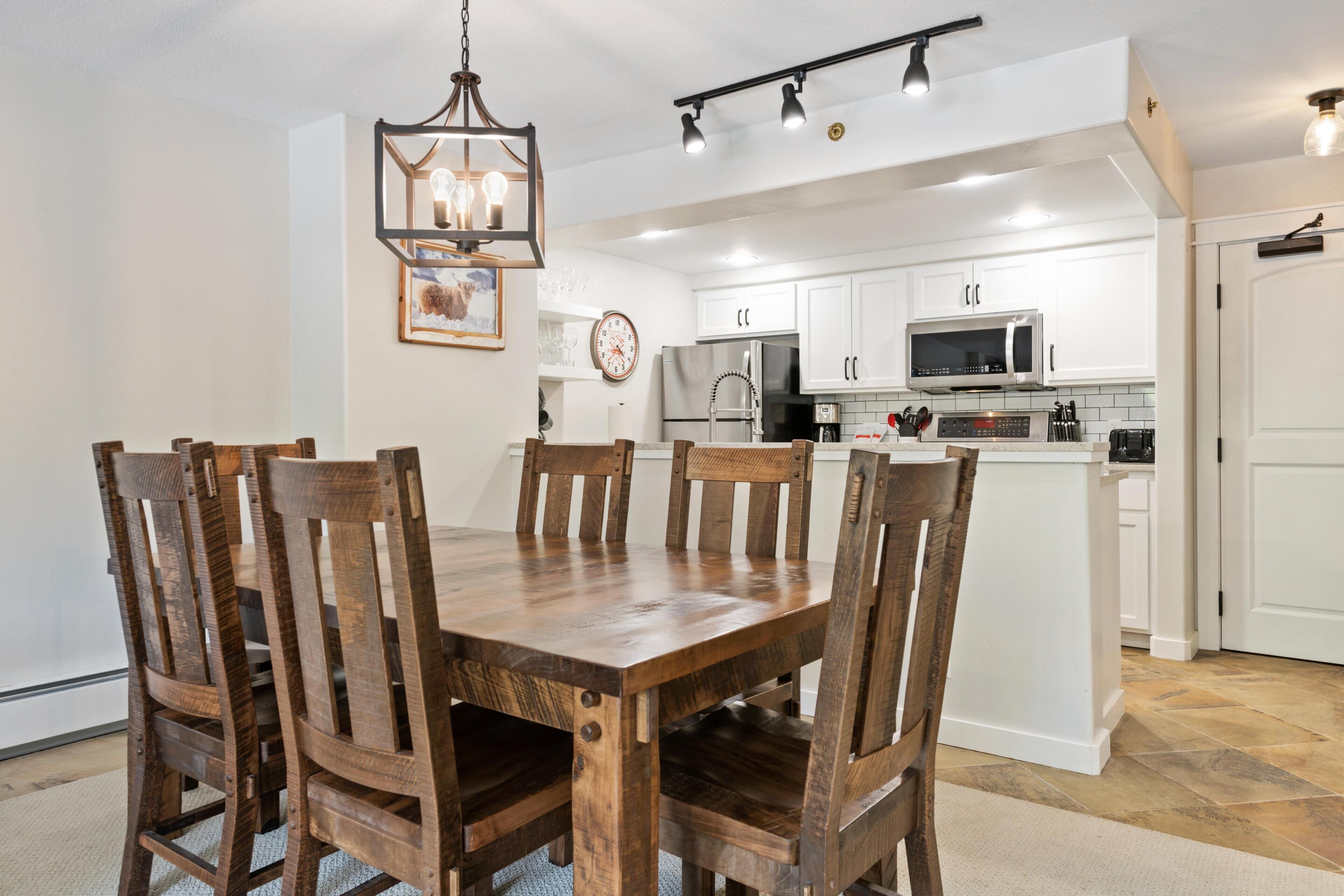 A dining area with a wooden table and chairs, a chandelier, and a kitchen with white cabinets and appliances.
