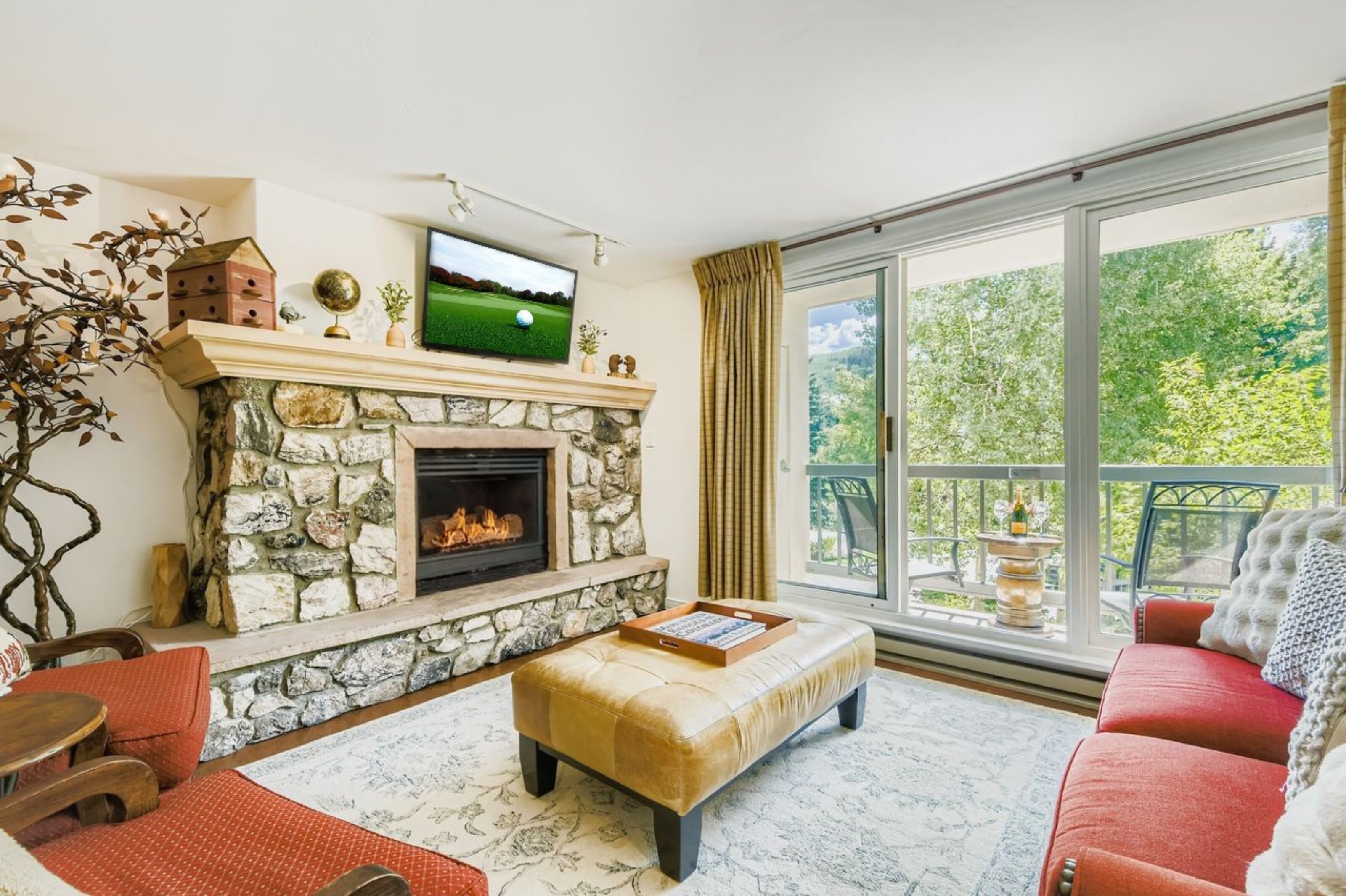 Un salón con chimenea de piedra, televisor, balcón con vistas a árboles y un sofá rojo.