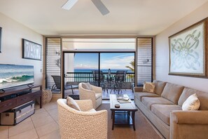 Ein Wohnzimmer mit Fernseher, Sofa und Sesseln, ein Balkon mit Tisch und Stühlen sowie Meerblick.