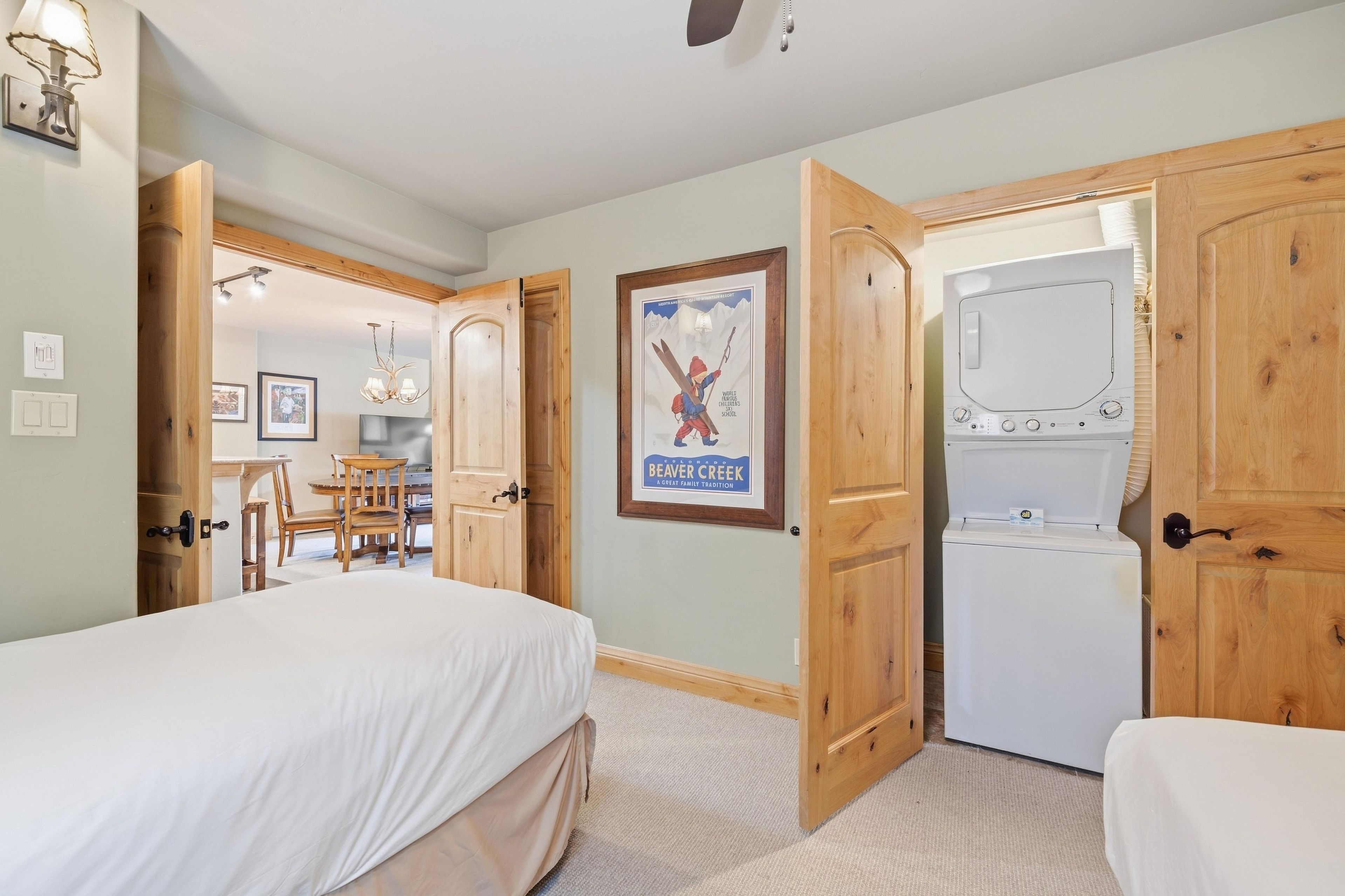 A bedroom with a bed, a desk, and a chair. There is a door leading to another room with a washing machine.