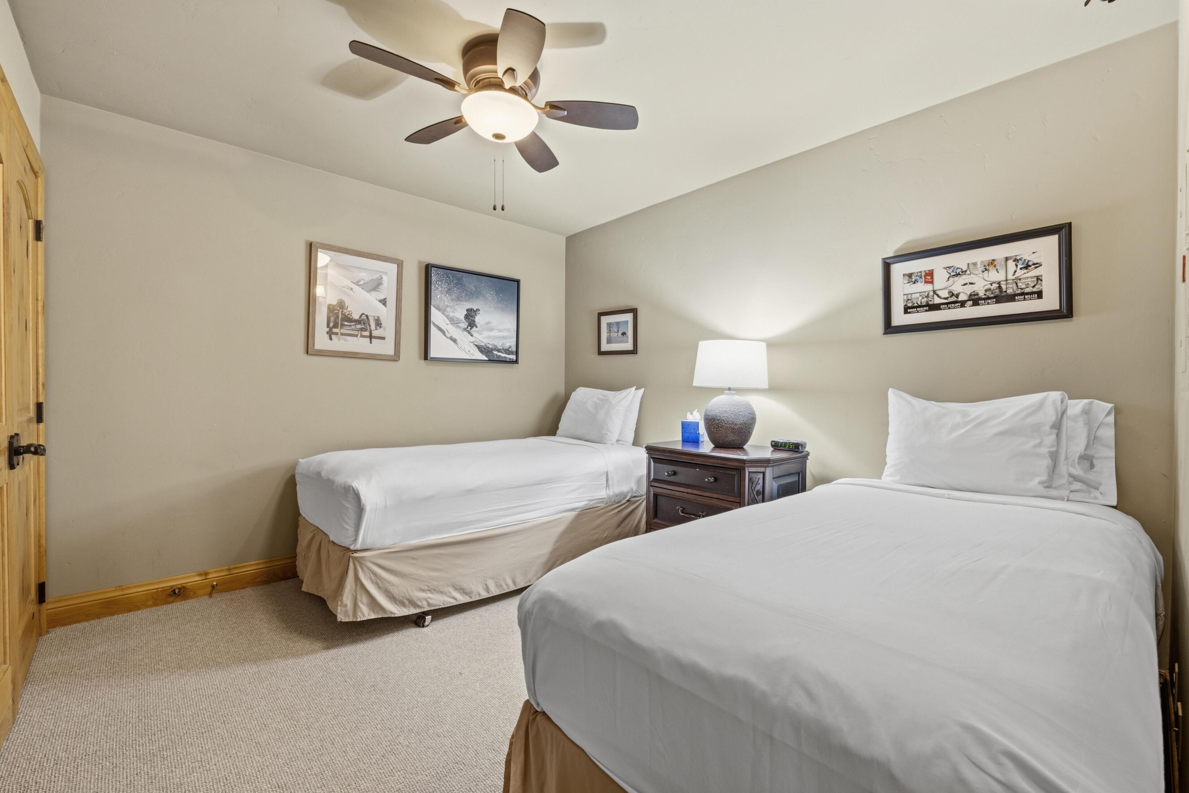 A hotel room with two beds, a ceiling fan, a nightstand, and framed pictures on the walls.