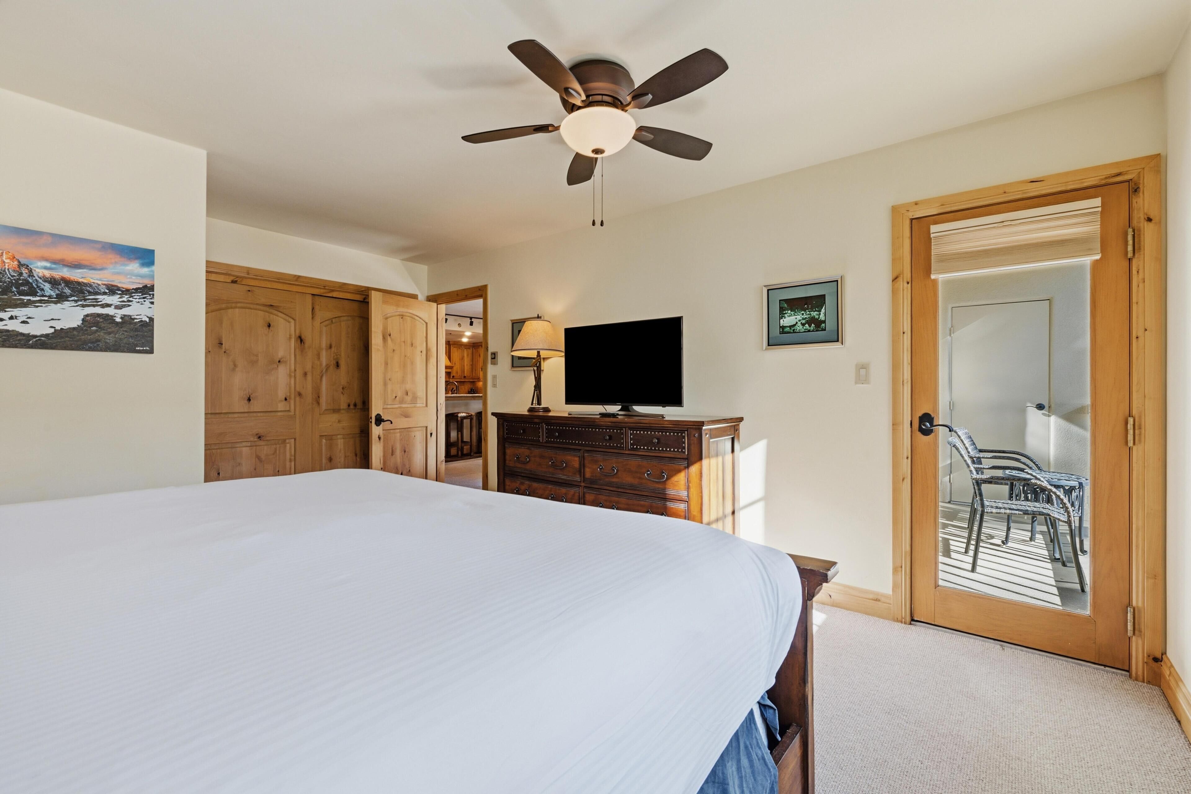 Quarto com cama, ventilador de teto, televisão, mobiliário de madeira e porta para área externa.