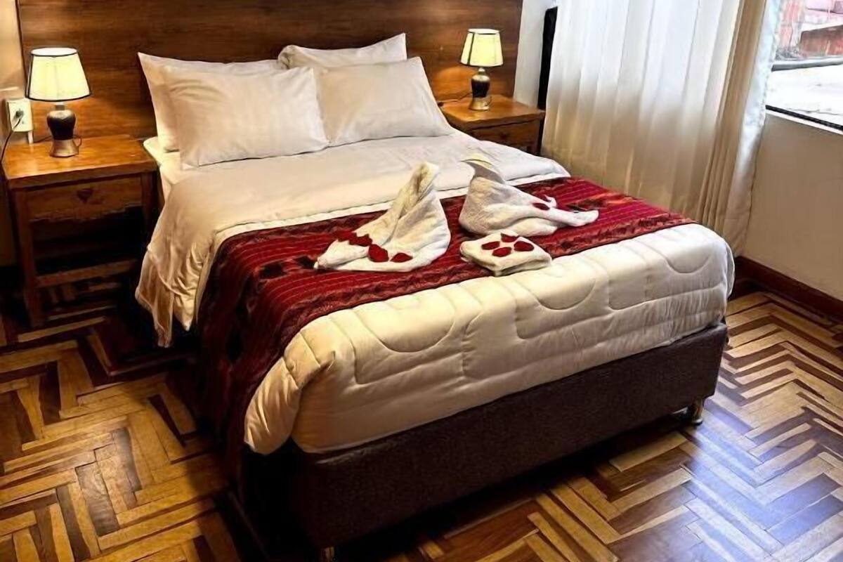 A hotel room with a wooden herringbone floor, a large bed with white bedding, two bedside lamps, and a framed picture on the wall.