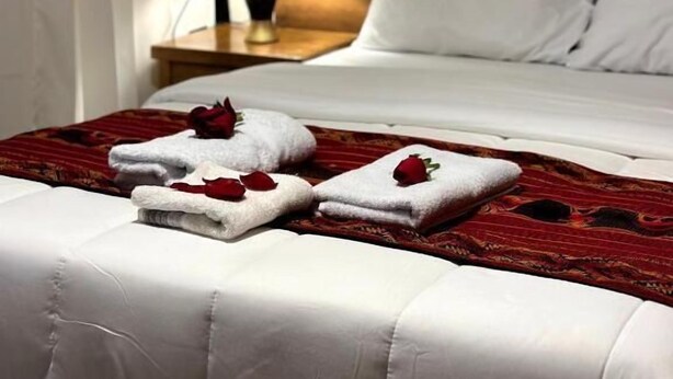 En snyggt bäddad säng med vita lakan och röda rosenblad, en sänggavel i trä, en sänglampa och vita gardiner.
