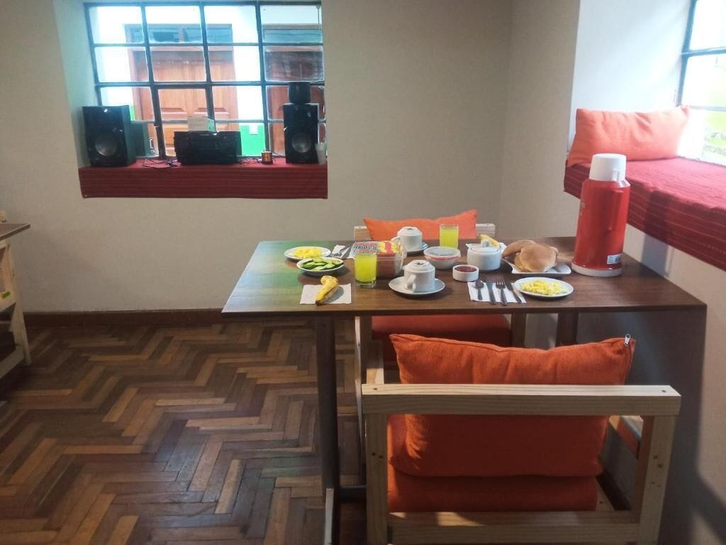 Un'area pranzo con un tavolo apparecchiato per la colazione, una finestra con degli altoparlanti e un letto con una coperta rossa.