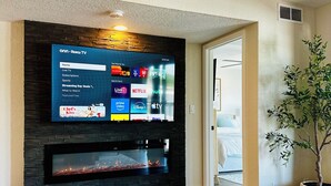 Ein Wohnzimmer mit einem an einer schwarzen Wand montierten Flachbildfernseher, einem Kamin darunter, Holzboden und einem Blumentopf.