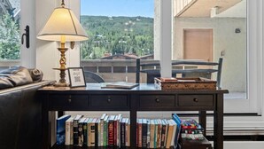Een kamer met een bureau, boeken, een lamp en uitzicht op een bos door grote ramen.