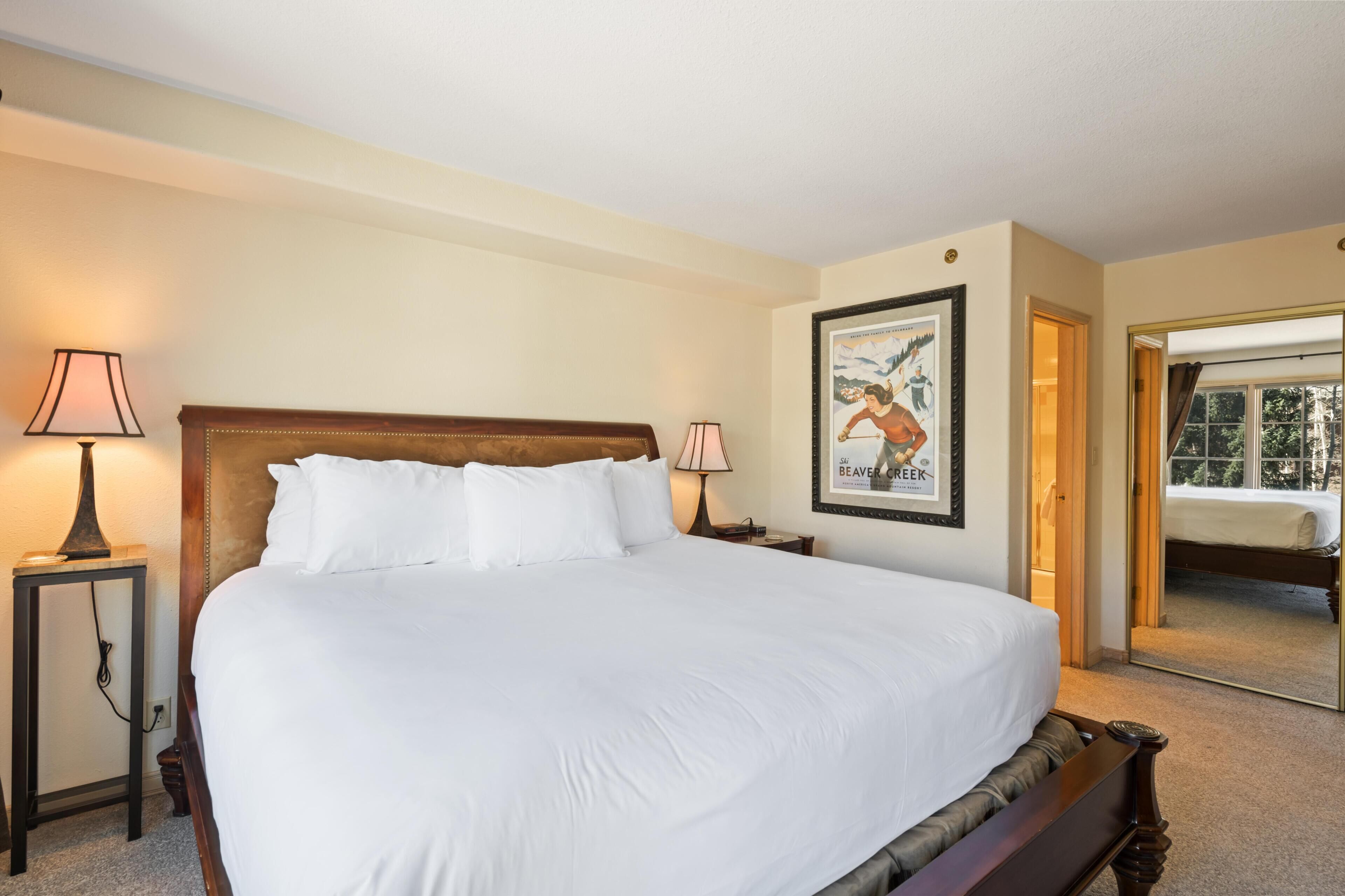 A neatly made bed in a hotel room with a headboard, two bedside lamps, a framed picture on the wall, and a mirror reflecting the room.