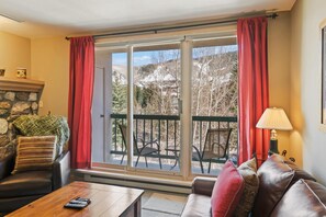 A cozy living room with a stone fireplace, a leather sofa, a wooden coffee table, and a view of a snowy landscape through large windows. - Borders Lodge L304 2BD Beaver Creek Ski-in, Ski-out Condo (Avon)
