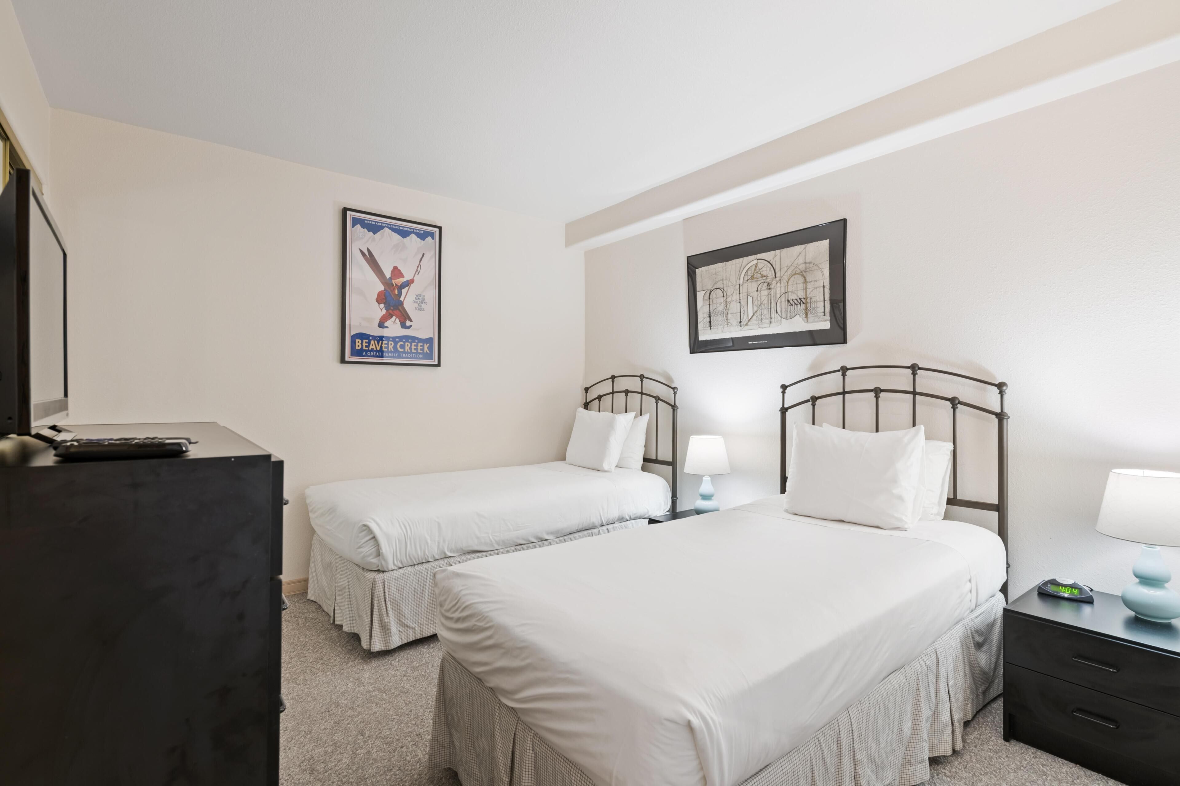 Habitación de hotel con dos camas, un escritorio, un aparador, un cuadro enmarcado y un cartel en la pared.
