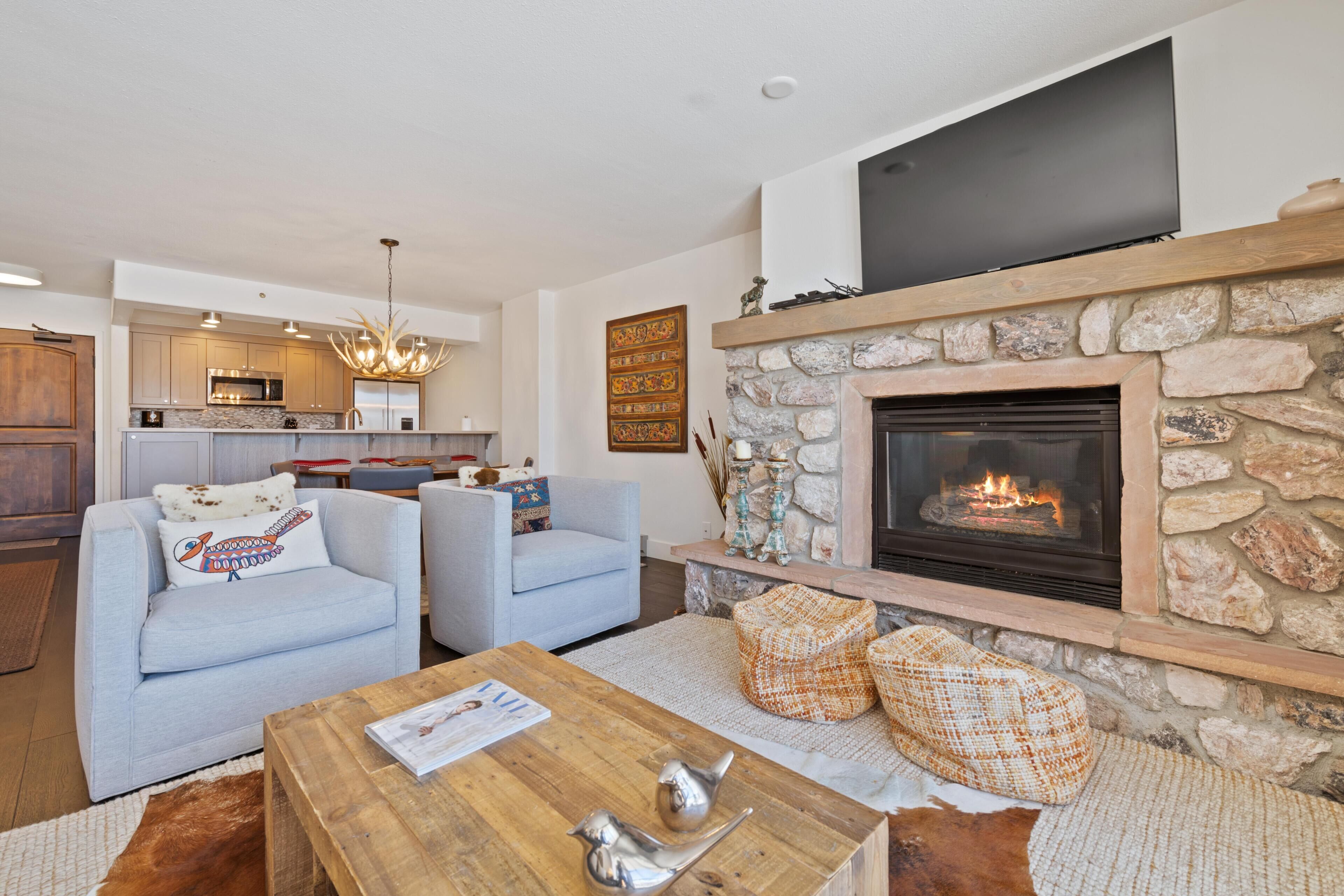 A modern living room with a fireplace, a kitchen in the background, and a seating area with a coffee table.