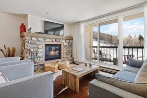 A cozy living room with a stone fireplace, comfortable seating, and a large window offering a view of the outdoors. - Borders Lodge L305 2BD Beaver Creek Ski-in, Ski-out Condo (Avon)