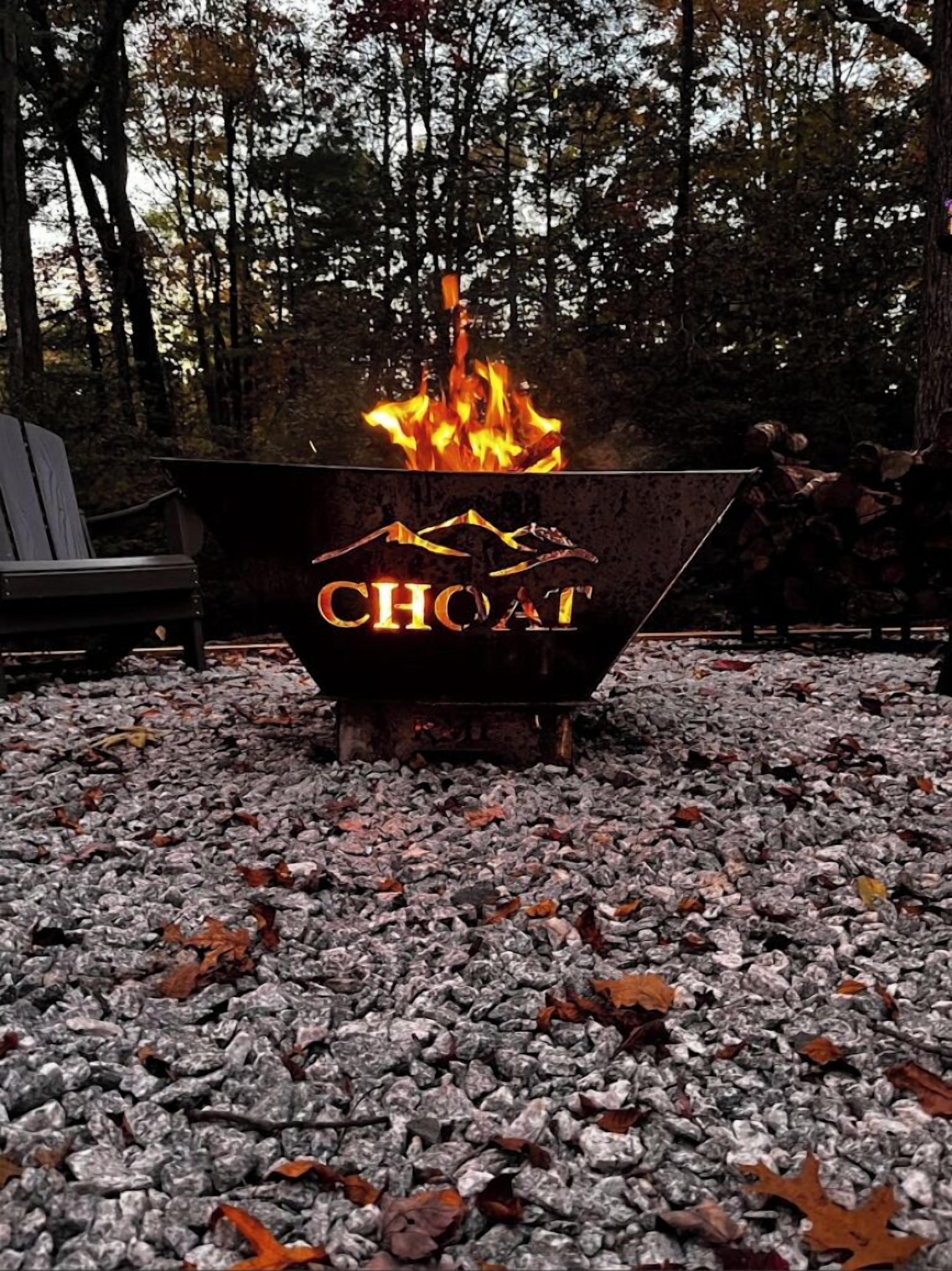 Ein Lagerfeuer mit einem 'CHOAT'-Schild, umgeben von Kies und Bäumen.