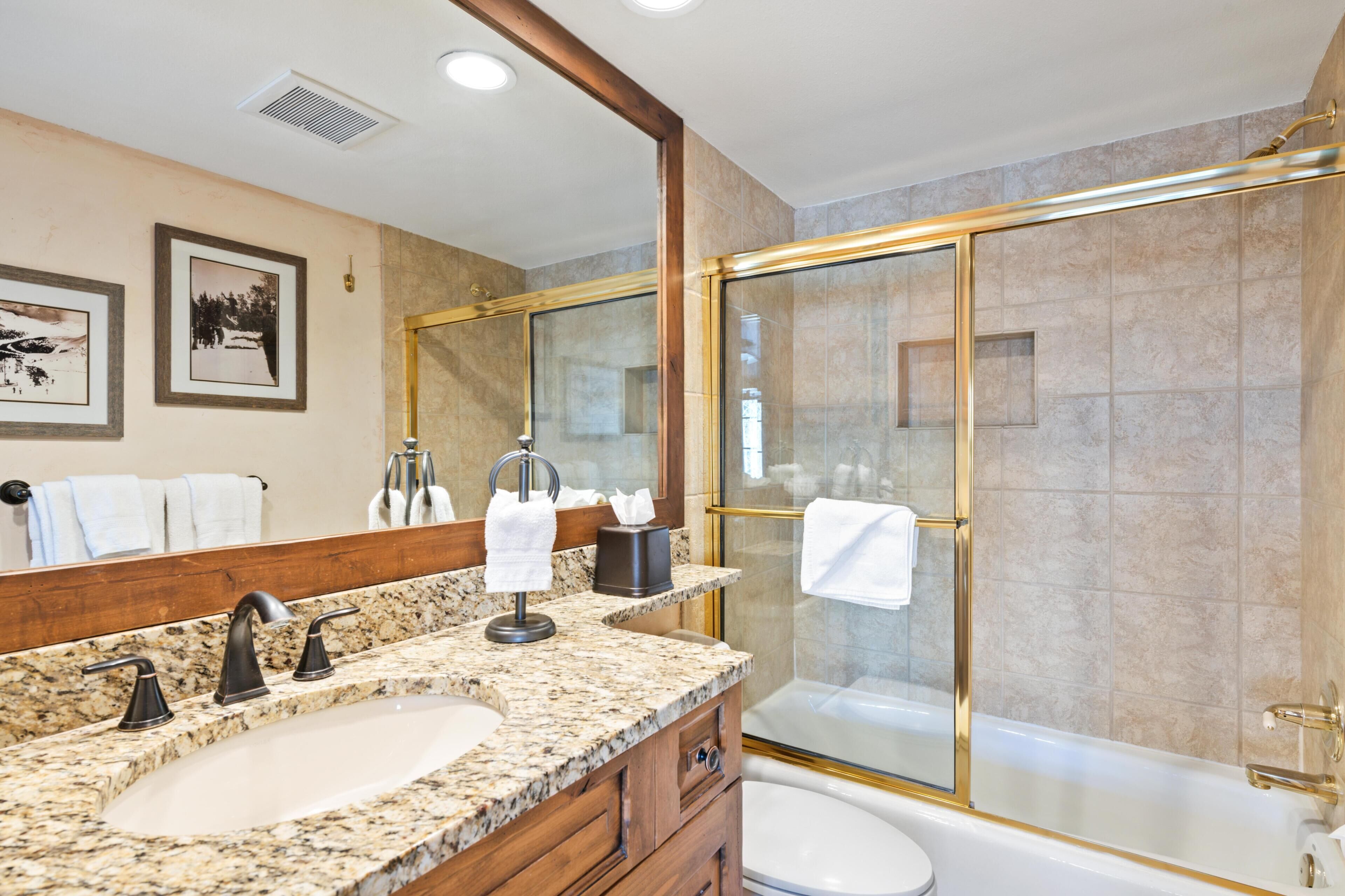 Un baño con encimera de granito, lavabo, espejo, ducha con puertas de cristal y retrete.