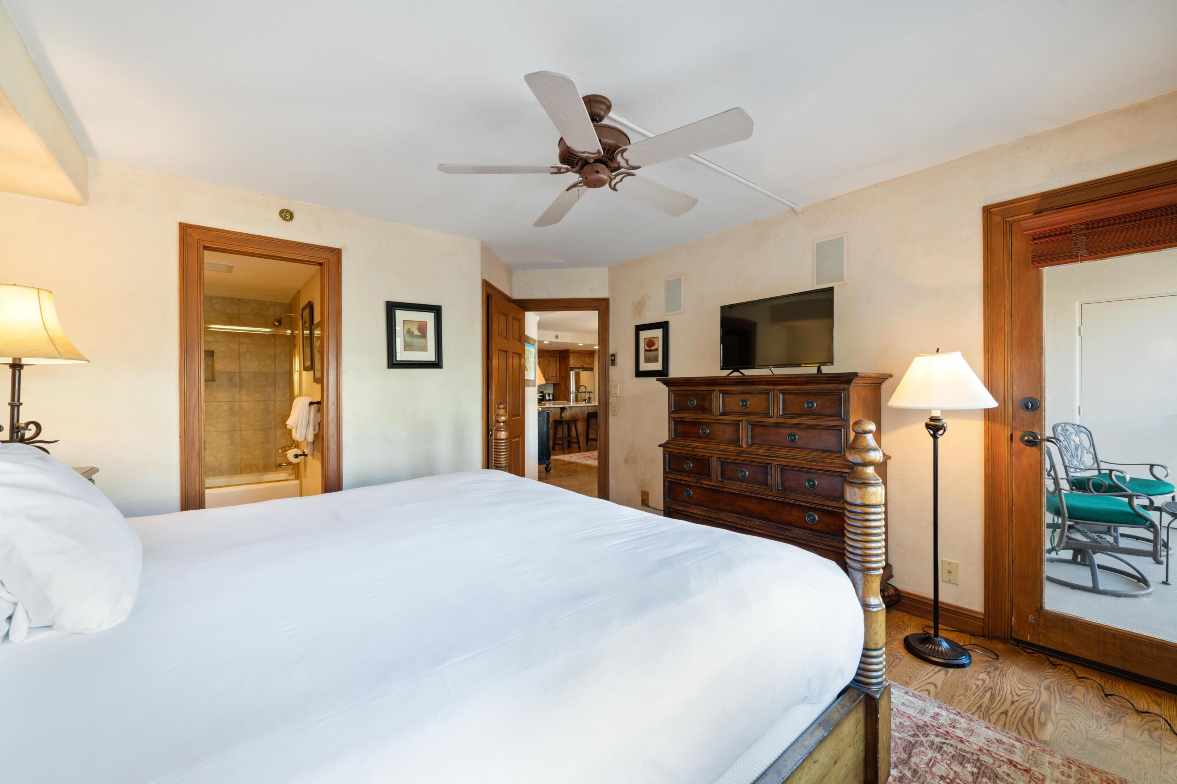 A hotel room with a bed, a ceiling fan, a dresser, a television, and a door leading to a bathroom.