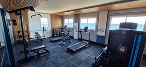 Ginásio com diversos equipamentos de exercício, esteira e vista para o mar através de grandes janelas.