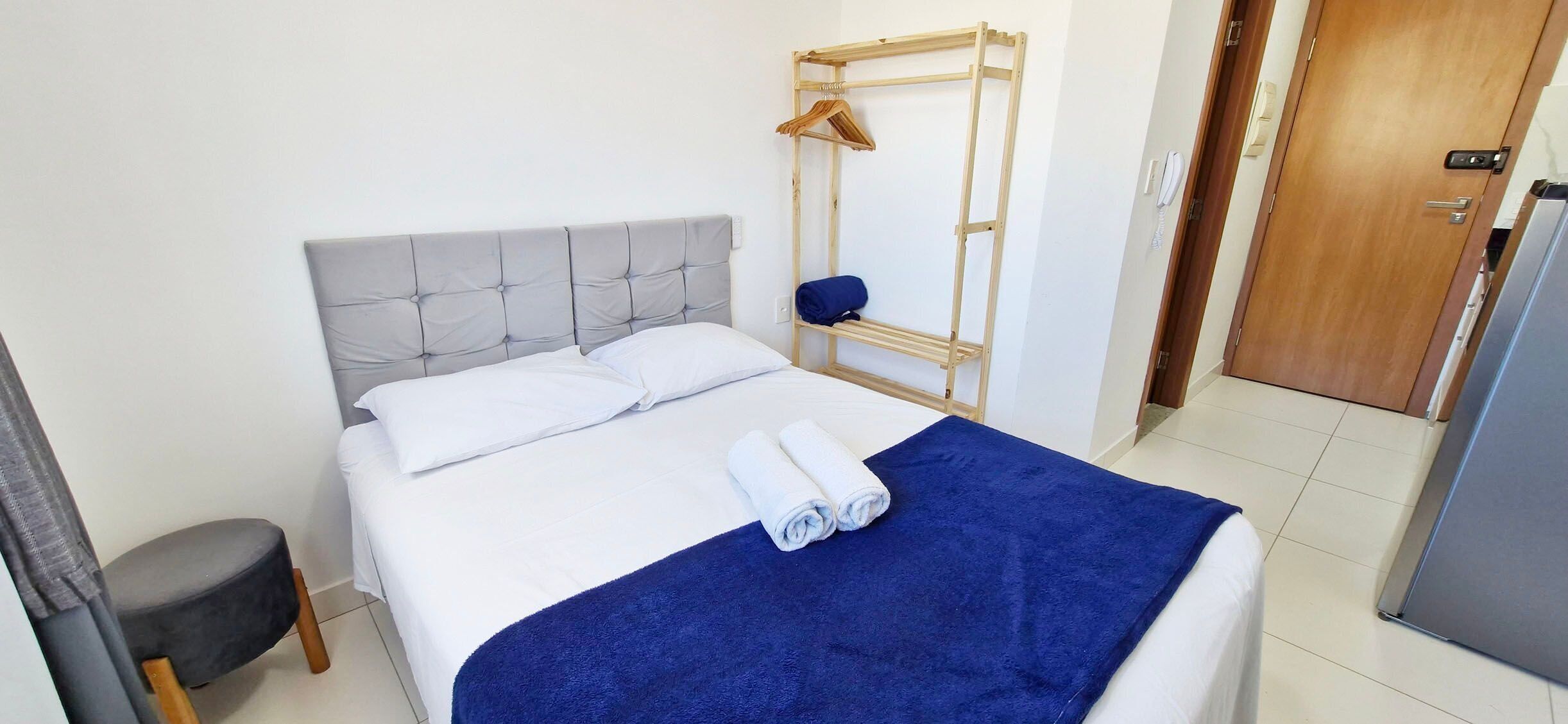 Una cama con cabecera gris tapizada, una manta azul y una toalla blanca enrollada. Se observa un armario de madera con una barra para colgar ropa y un taburete gris.
