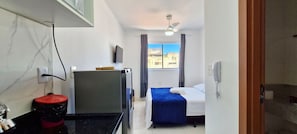 Quarto de hotel com cama, televisão, escrivaninha, lixeira e janela com cortinas.