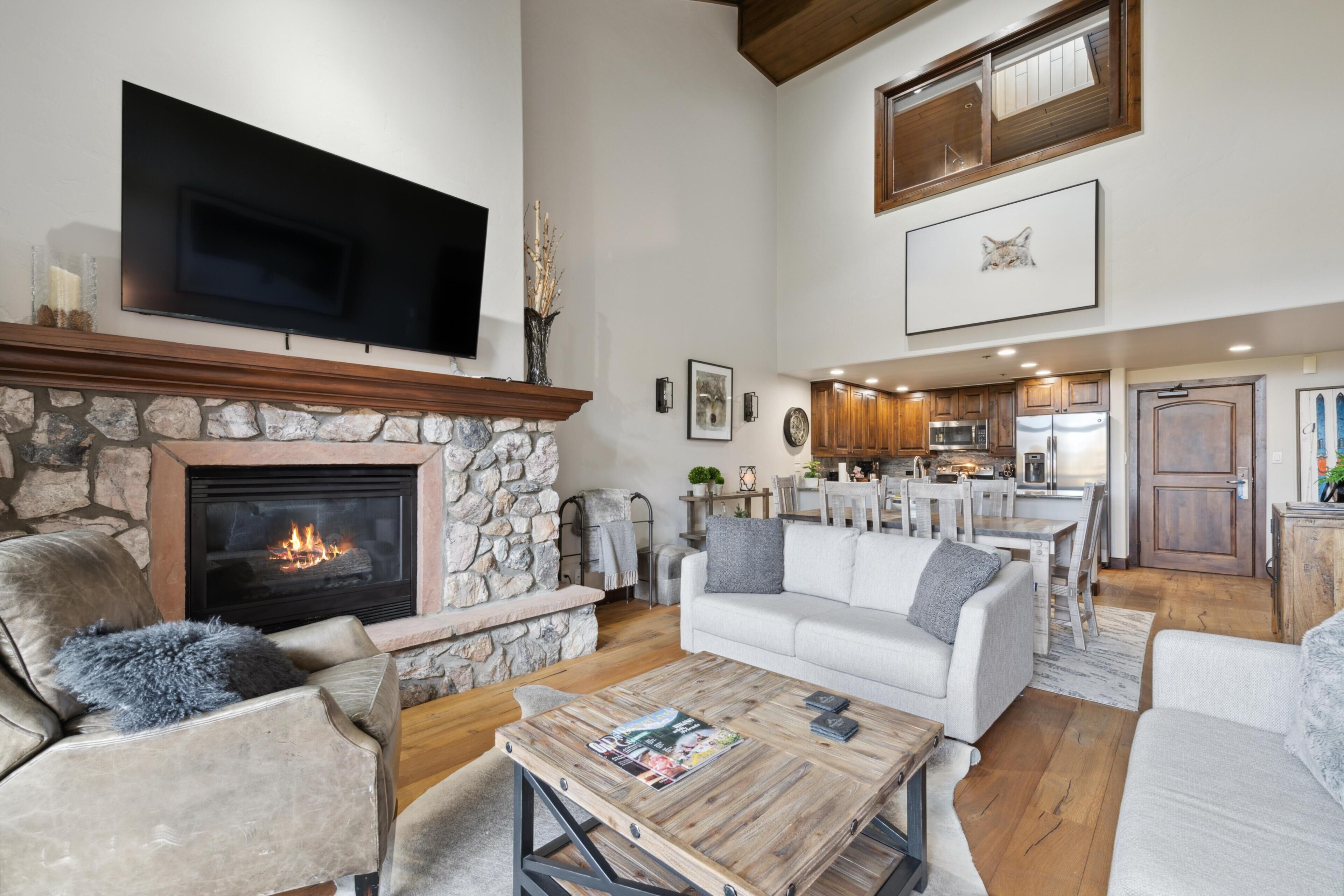 A spacious living area with a stone fireplace, a flat-screen TV, a dining area, and a kitchen with wooden cabinets.