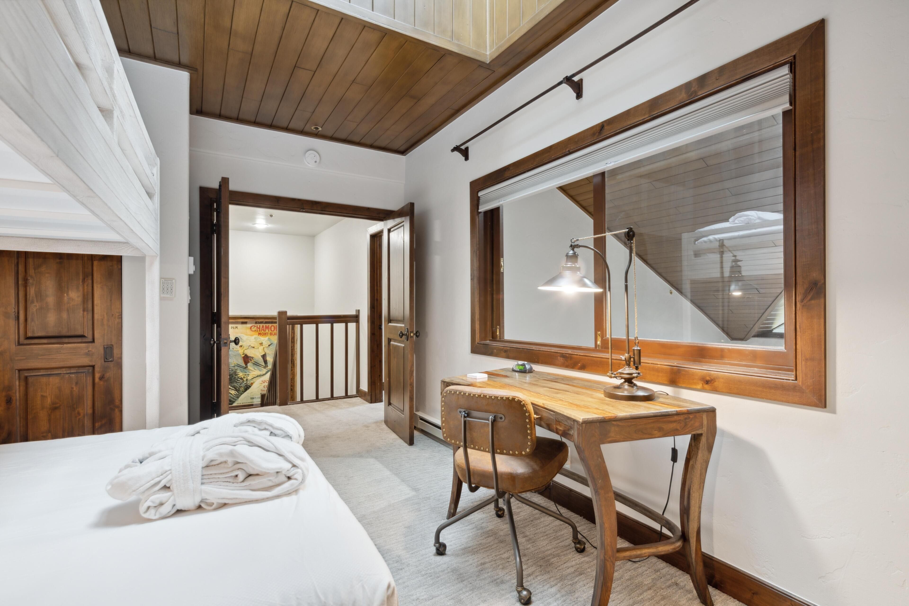 A bedroom with a wooden desk, a chair, a bed with a rolled-up towel, a large mirror, and a window.
