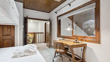 A bedroom with a wooden desk, a chair, a bed with a rolled-up towel, a large mirror, and a window.