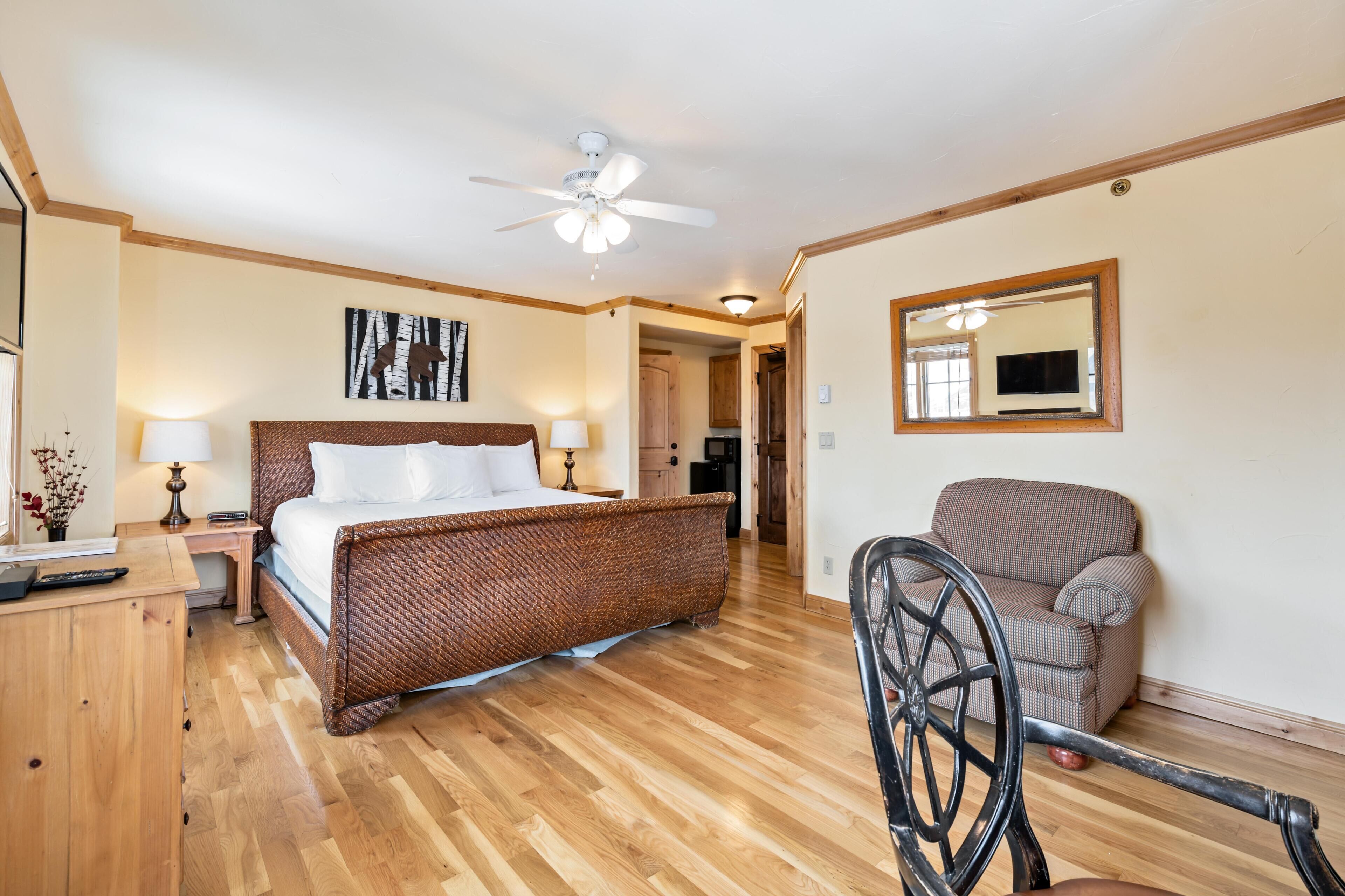 A bedroom with a wooden floor, a bed with white linens, a ceiling fan, a chair, and a mirror on the wall.