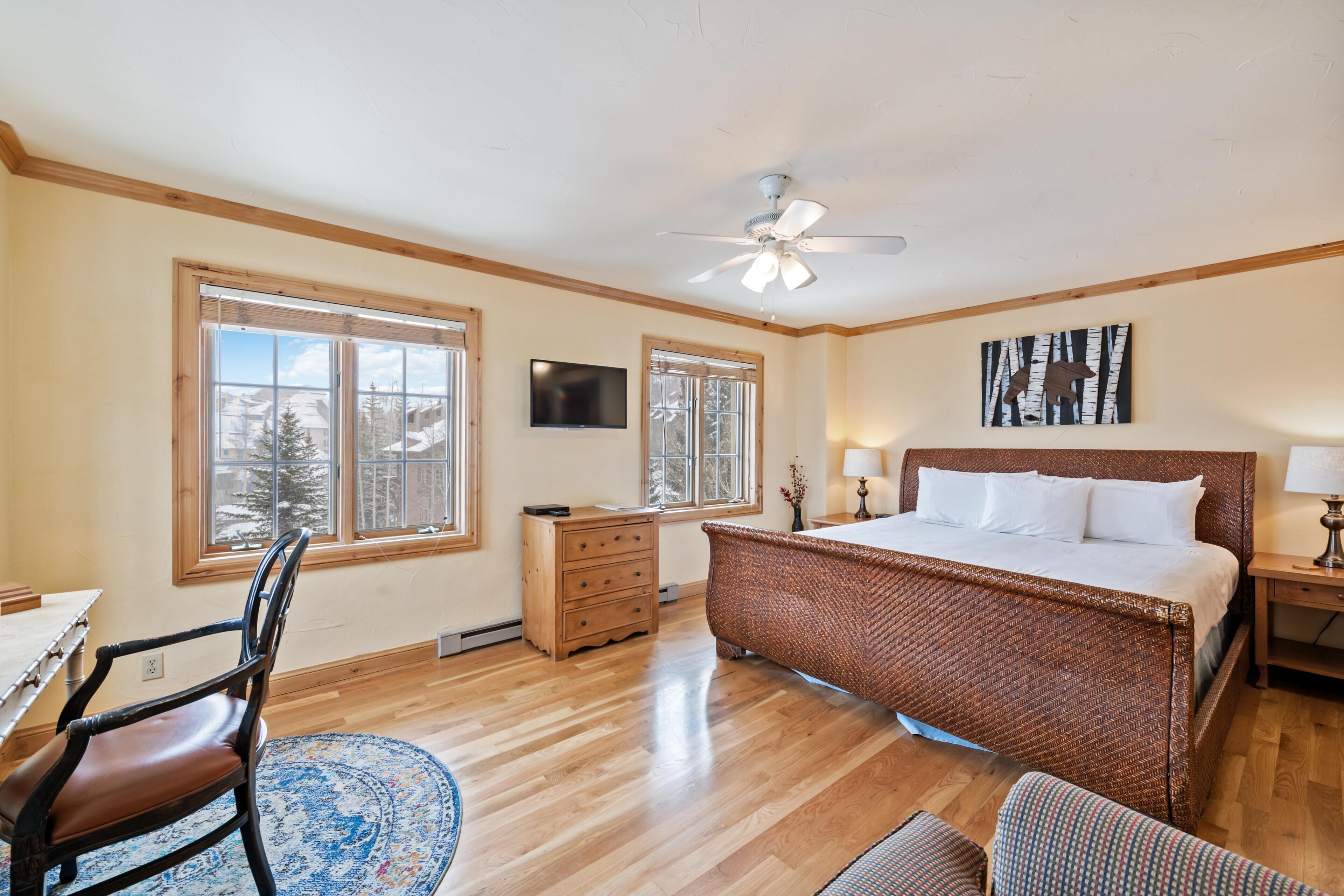 A bedroom with a large bed, wooden floor, a ceiling fan, and a window with a view of trees.