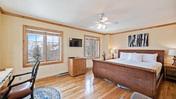 A bedroom with a large bed, wooden floor, a ceiling fan, and a window with a view of trees.