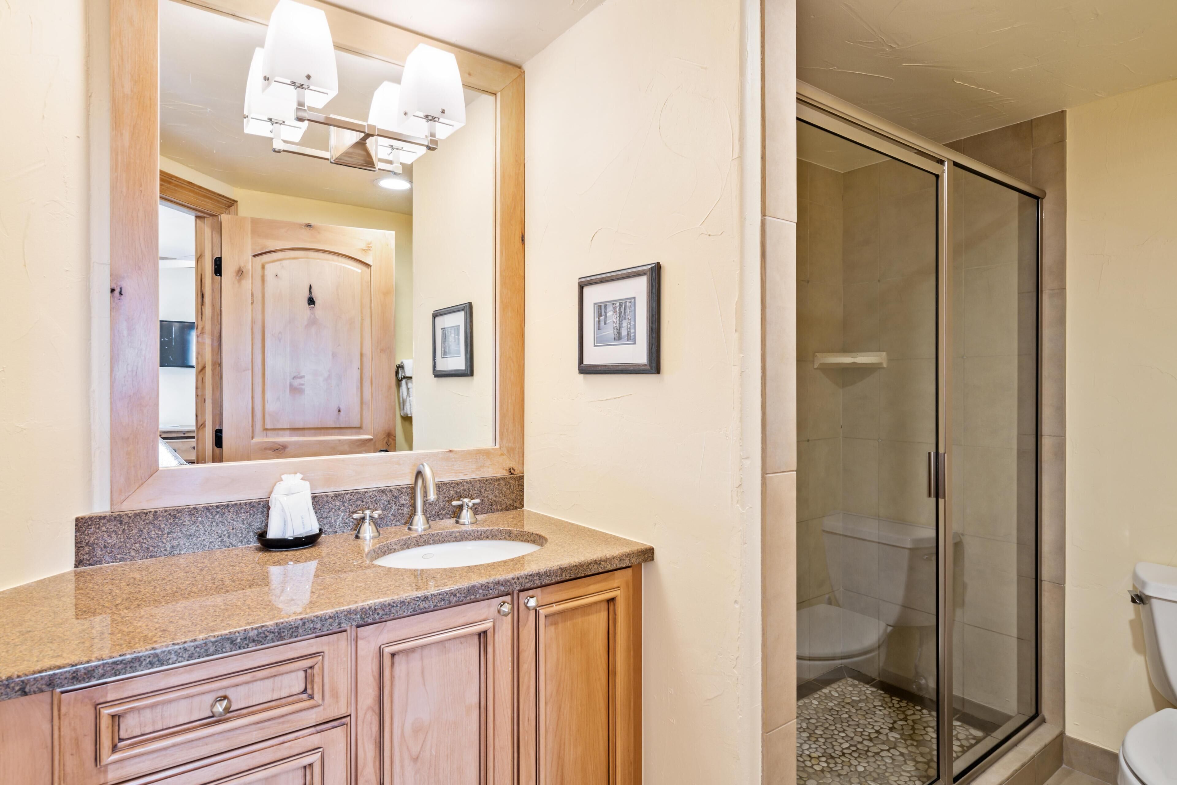 A bathroom with a granite countertop sink, wooden cabinets, a large mirror, and a glass-enclosed shower.