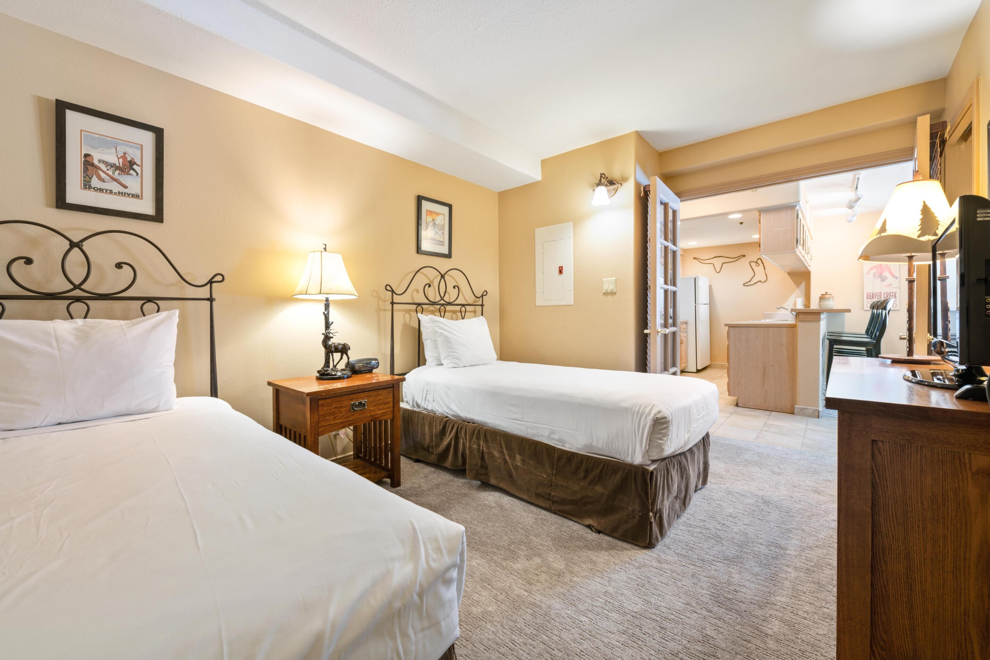 A hotel room with two beds, a desk, a lamp, a dresser, and a kitchenette.