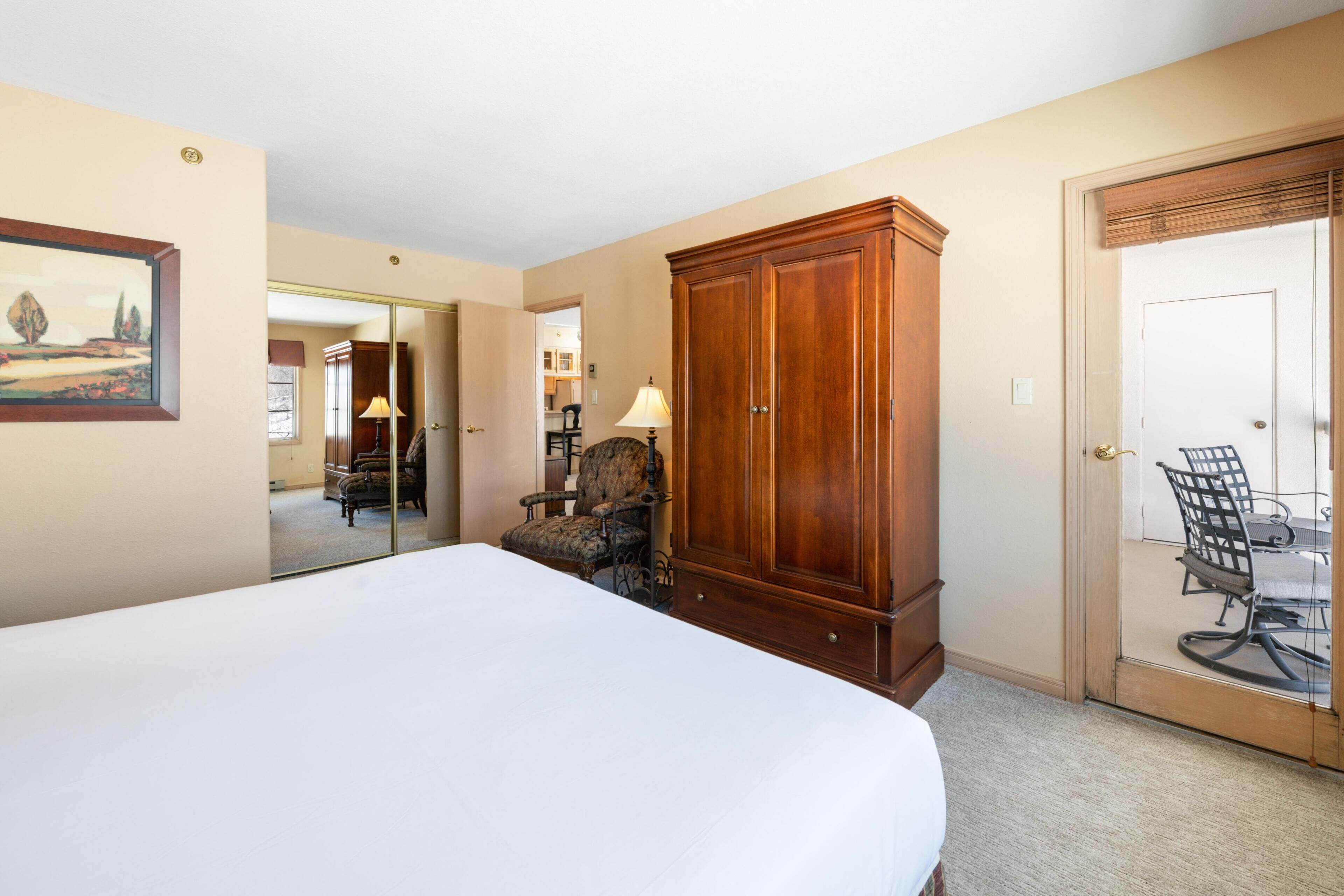 Habitación de hotel con una cama grande, un armario de madera, una silla y una mesa con sillas afuera de la puerta.