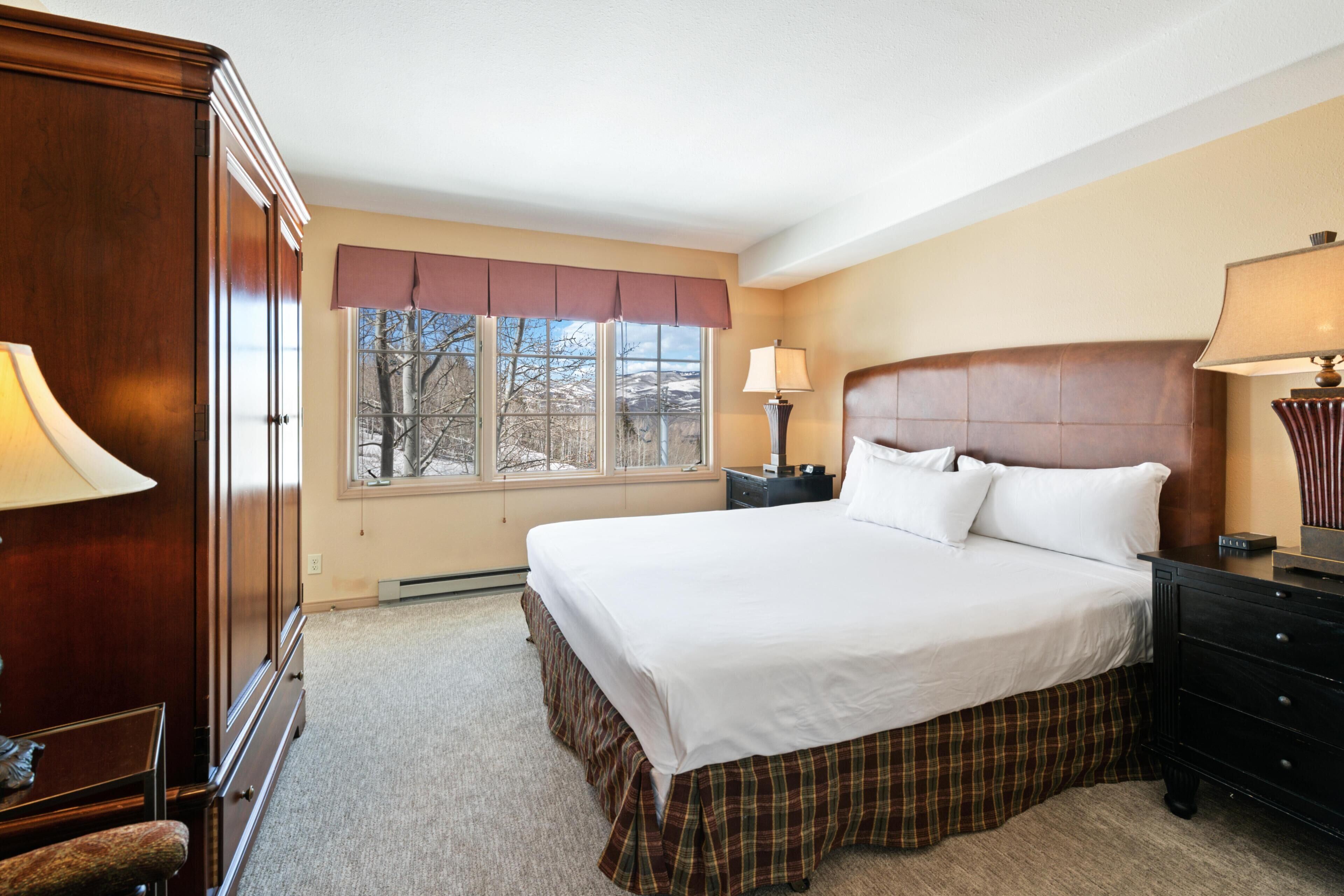 A bedroom with a large bed, a nightstand, a dresser, and a window with a view of trees.