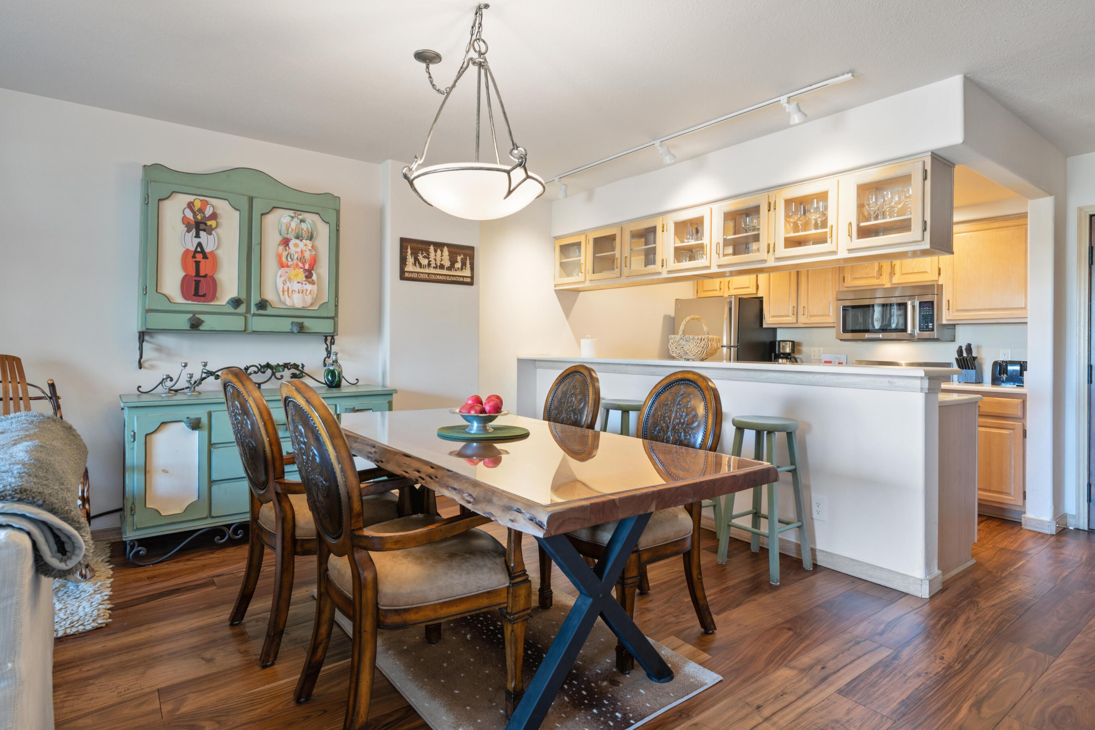 Un comedor con una mesa de madera, sillas y una lámpara de araña. Hay una zona de cocina con armarios y un microondas.