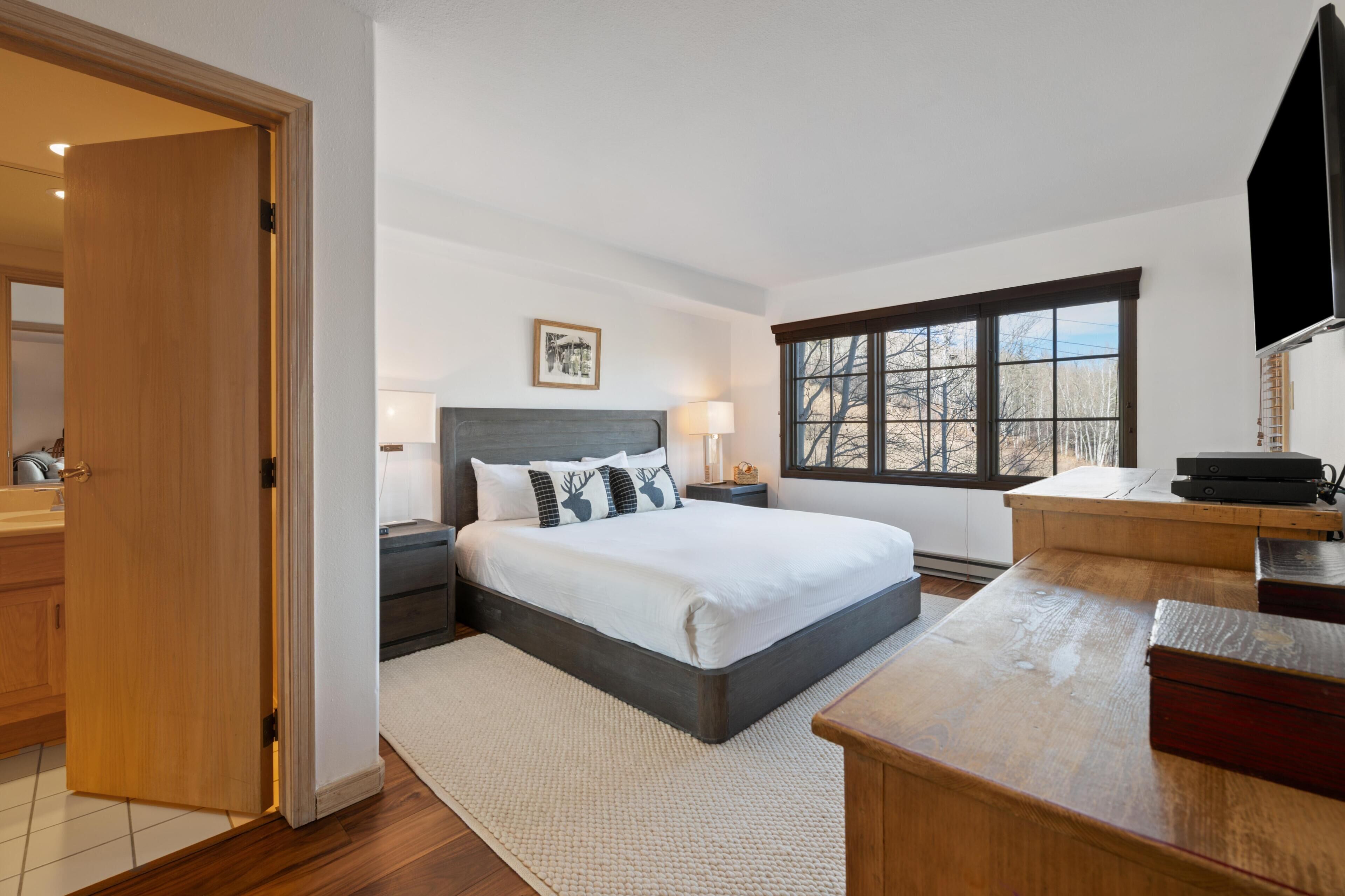 A bedroom with a bed, bedside table, lamp, window, and a TV.