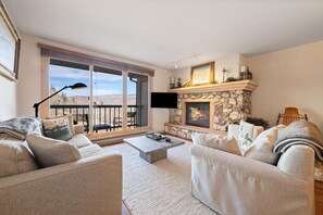 A living room with a stone fireplace, a large window, and a balcony. - Borders Lodge L403 2BD Beaver Creek Ski-in, Ski-out Condo (Avon)