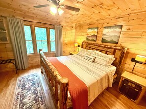 Une chambre à coucher dans une cabane en bois, avec un lit, une table de chevet, une lampe, une fenêtre avec des rideaux et un ventilateur de plafond.